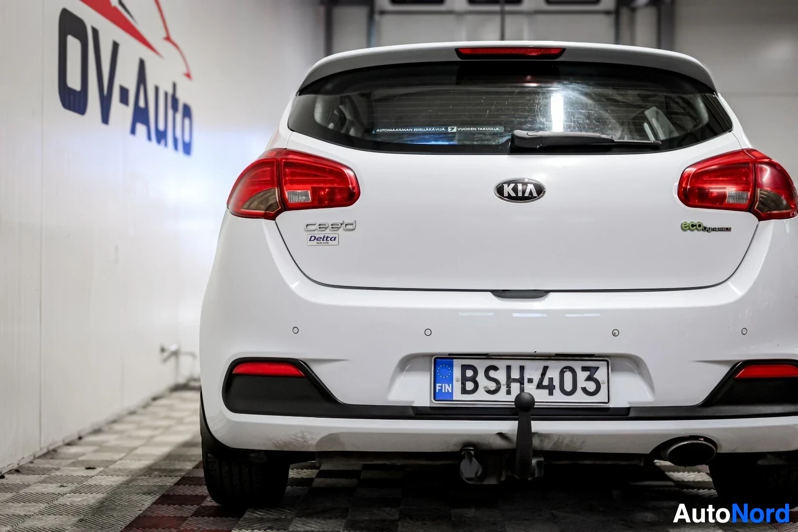 Takaa: Valkoisen hatchback-auton peräosa sisätiloissa. Autossa on vetokoukku, pakoputki näkyvissä oikealla, ja rekisterikilpi. Takapuskurissa on kolme pyöreää