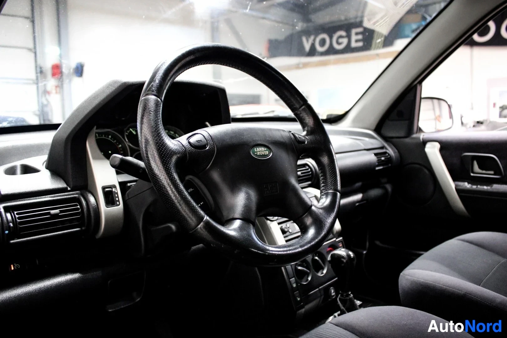 Ohjaamo: Kuvakulma auton ohjaamosta, joka näyttää ohjauspyörän, kojelaudan ja osan etuistuimesta. Ohjauspyörässä on "LAND ROVER" -logo ja useita painikkeita. Kojelaudass