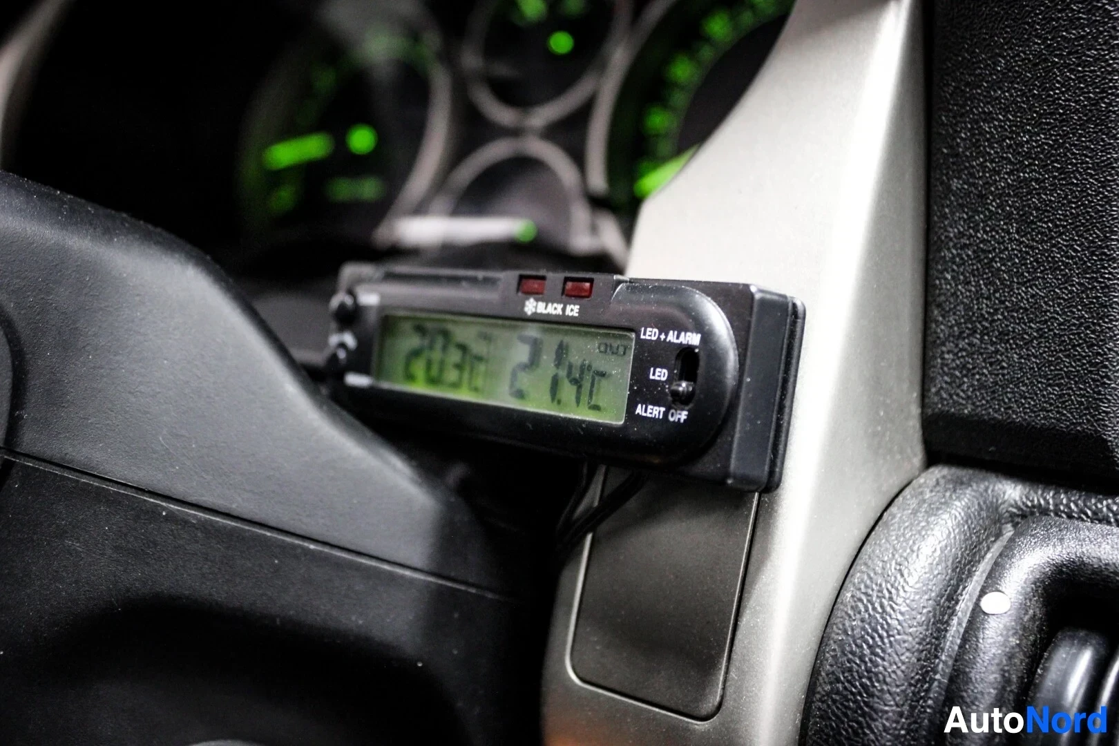 Yksityiskohta: Lähikuva autoon jälkiasennetusta Black Ice -merkkisestä ulkolämpötilan ja ajan näytöstä, joka näyttää lukemat 20:38 ja 21.4C. Taustalla näkyy ajoneuvon mittaris