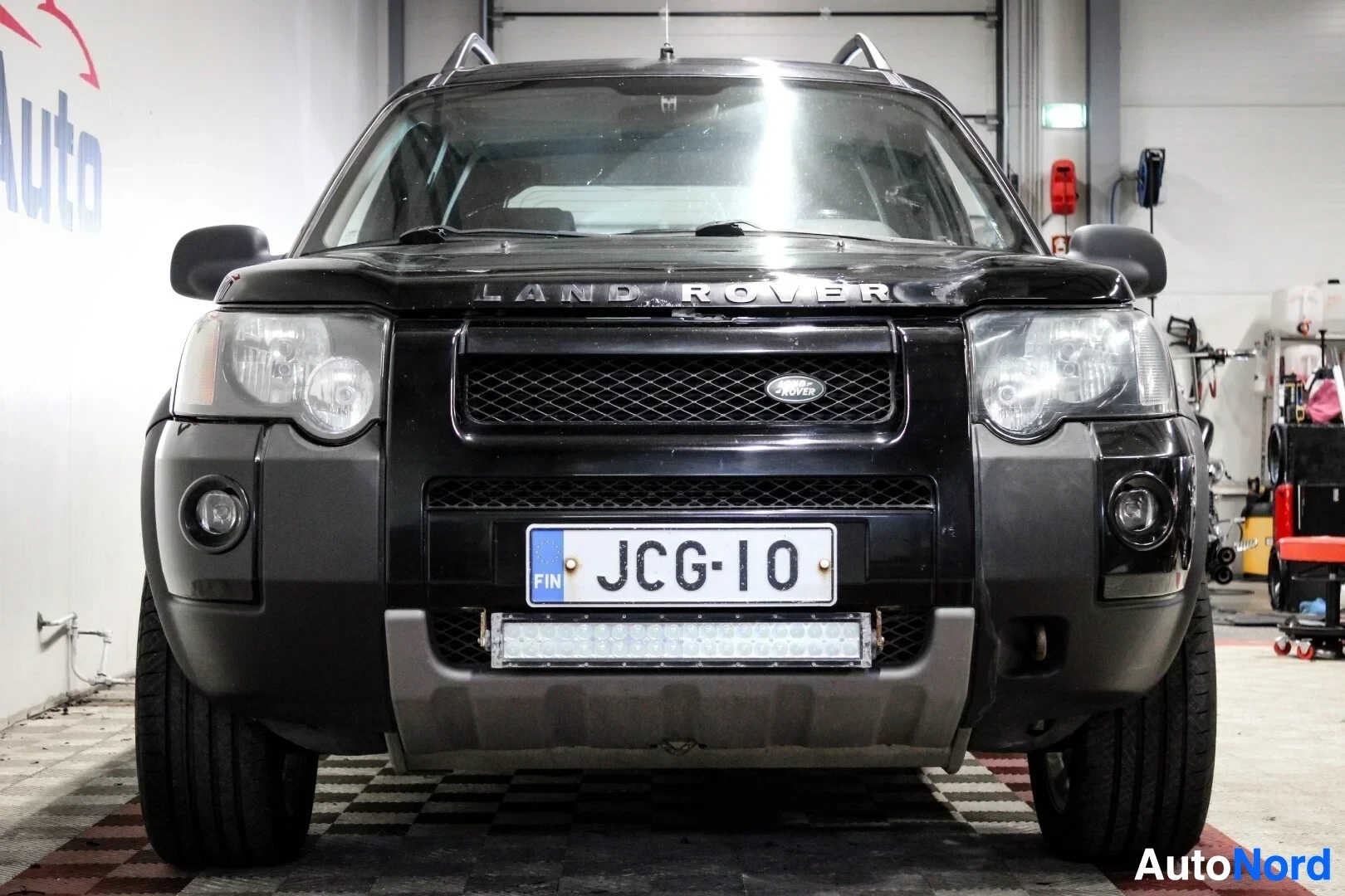 Edestä: Land Rover -tekstillä varustetun mustan maastoauton etuosa. Ajoneuvossa on valkoinen rekisterikilpi "" ja puskurissa lisävalopaneeli