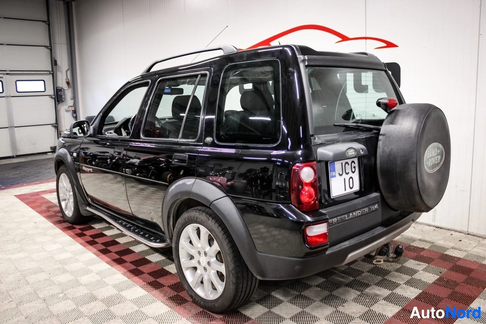 Takaviisto: Musta Land Rover Freelander pysäköitynä halliin. Ajoneuvon vasen kylki ja takaosa ovat näkyvissä. Takaluukussa on vararengas suojapeitteellä ja peräkoukku. Taka