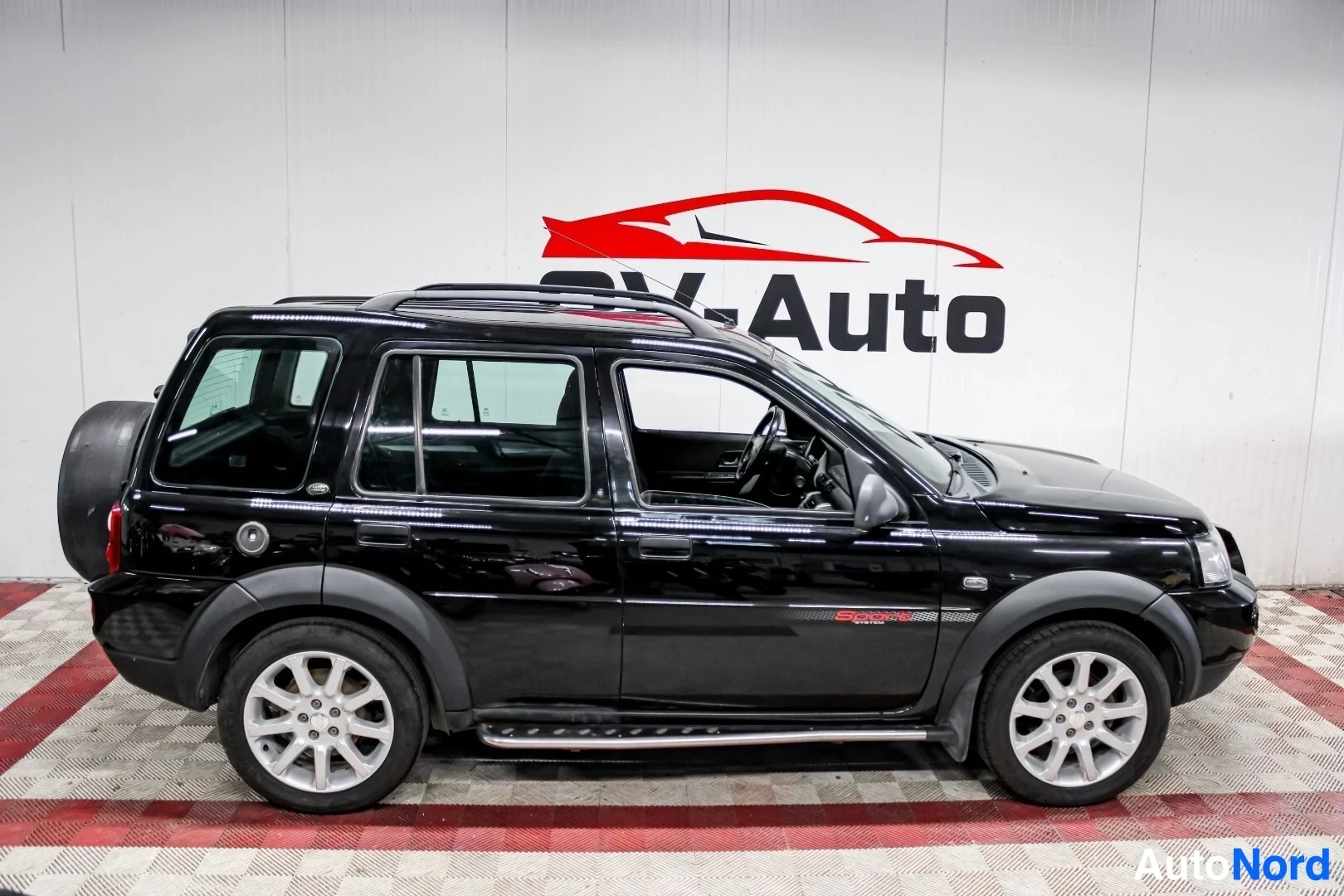 Sivulta: Musta Land Rover Freelander pysäköitynä halliin. Ajoneuvon oikea kylki on näkyvissä. Ajoneuvossa on kattokaiteet ja astinlaudat. Takaluukussa on vararengas suoj