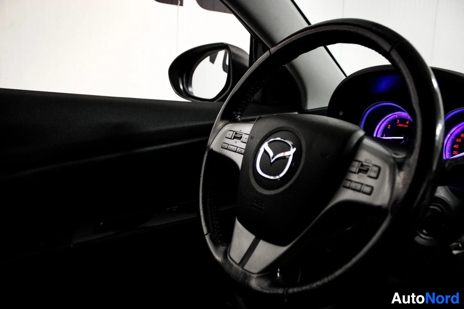 Ohjaamo: Lähikuva ohjauspyörästä ja mittaristosta. Ohjauspyörässä on Mazda-logo keskellä ja useita painikkeita. Mittaristo palaa violettina. Ikkunasta näkyy ajoneuvon si