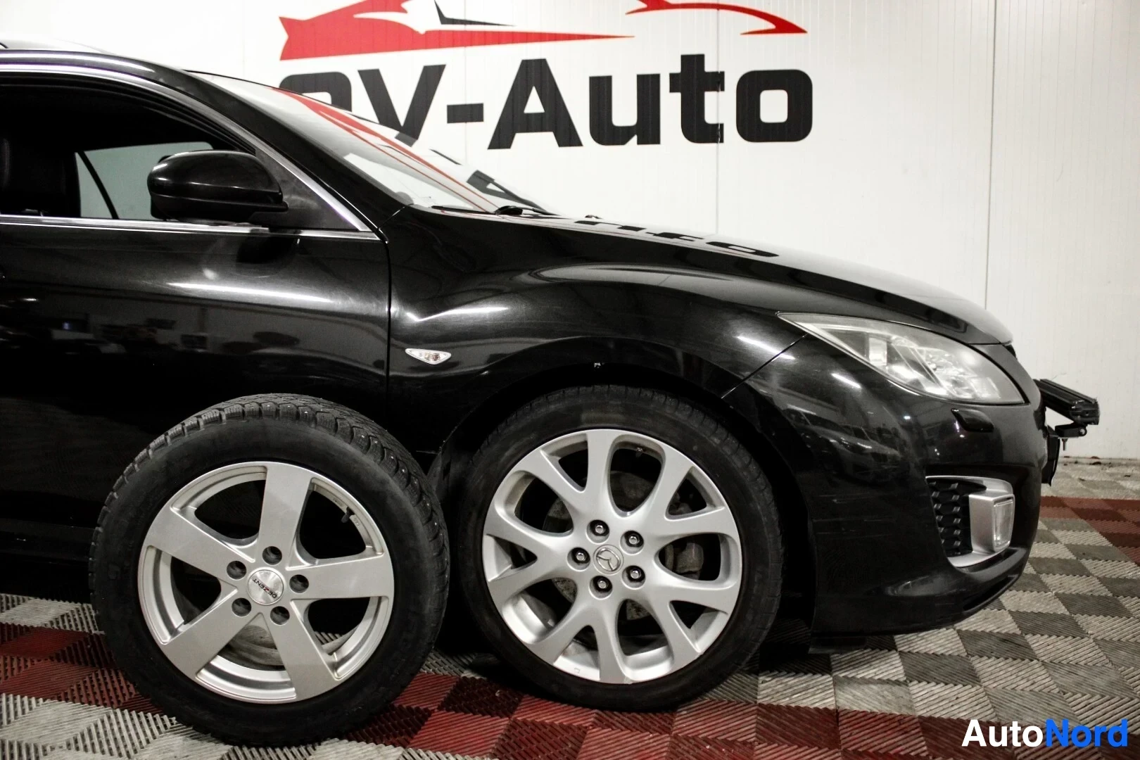 Etuviisto, nokka oikealle: Musta auto seisoo kuvassa edestä vasemmalta, edessä näkyy kaksi rengassarjaa, toinen on asennettu autoon ja toinen seisoo sen vieressä.