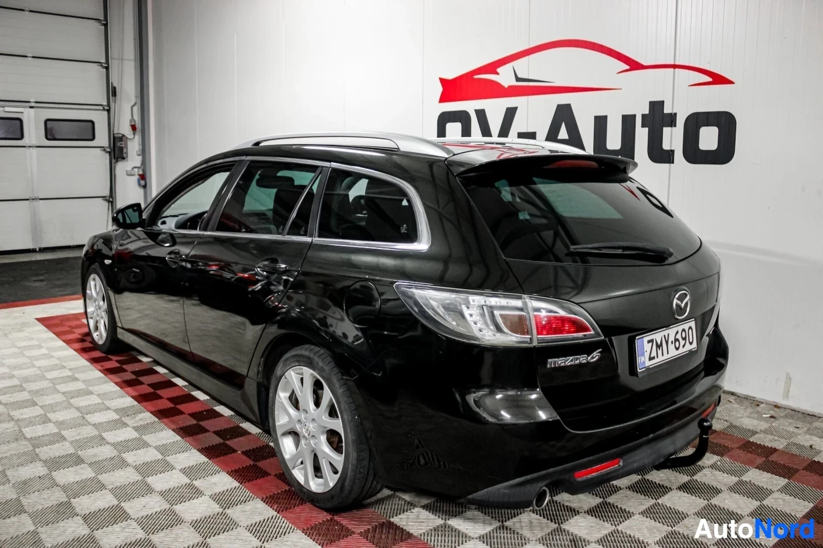 Takaviisto: Musta farmariauto takaa ja vasemmalta viistosti kuvattuna OV-Auto-logon edessä. Auton katolla on kattokiskot ja alla vaaleat kevytmetallivanteet. Takaluukun ala