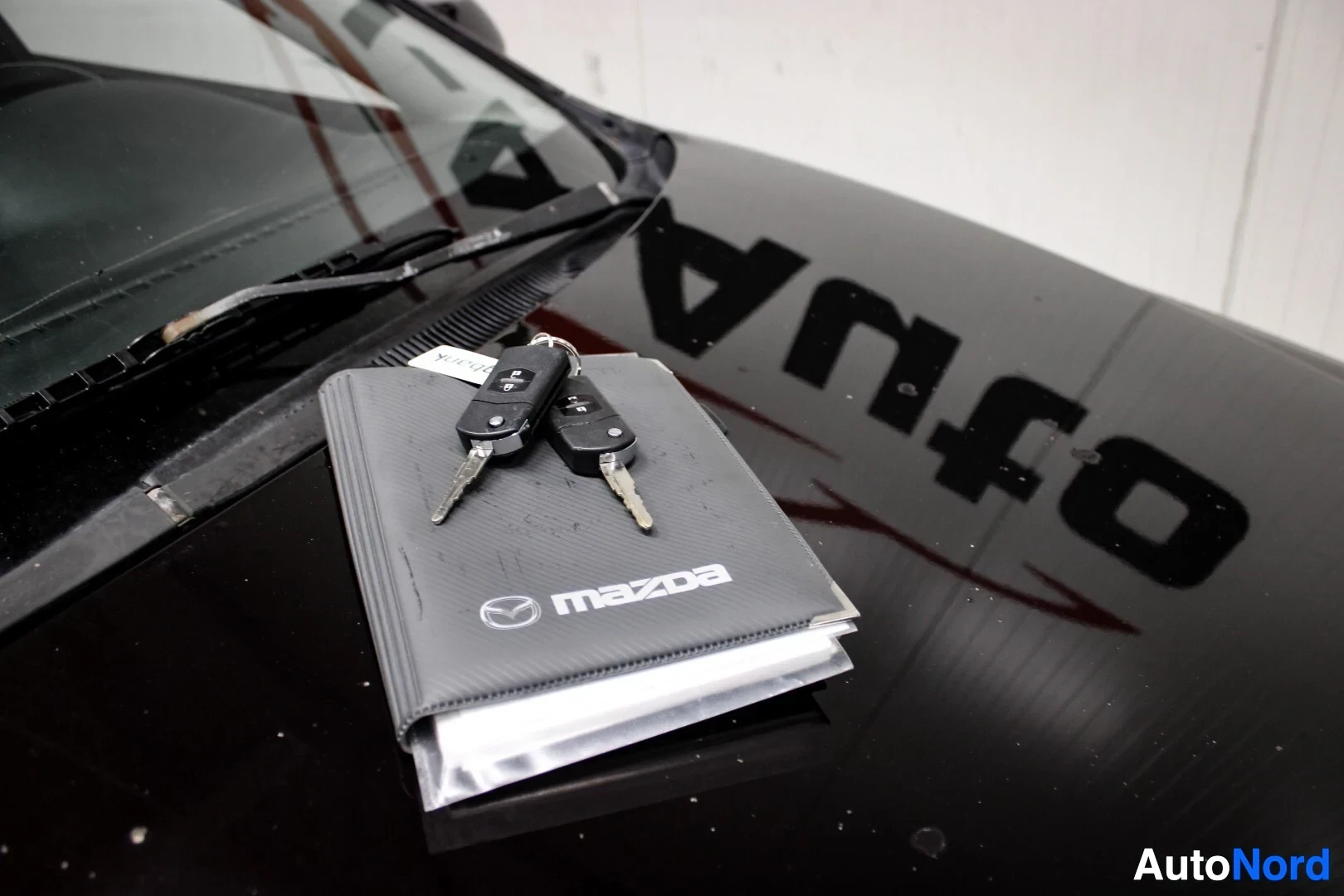 Yksityiskohta: Kaksi auton avainta ja ohjekirja "mazda"-logolla mustalla konepellillä, jossa heijastuu valkoista tekstiä.