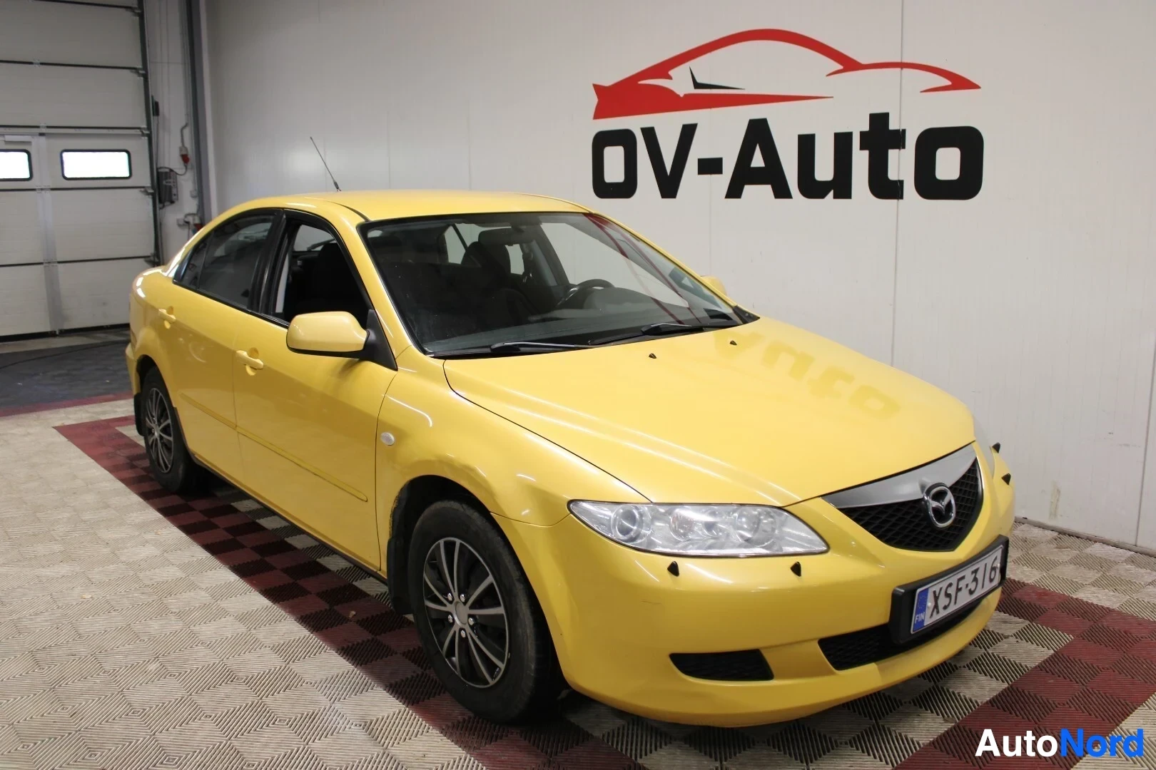 Etuviisto, nokka oikealle: Keltainen sedan auto kuvattuna etuviistosta vasemmalta, edessä rekisterikilpi "" ja musta "OV-Auto"-logo seinällä