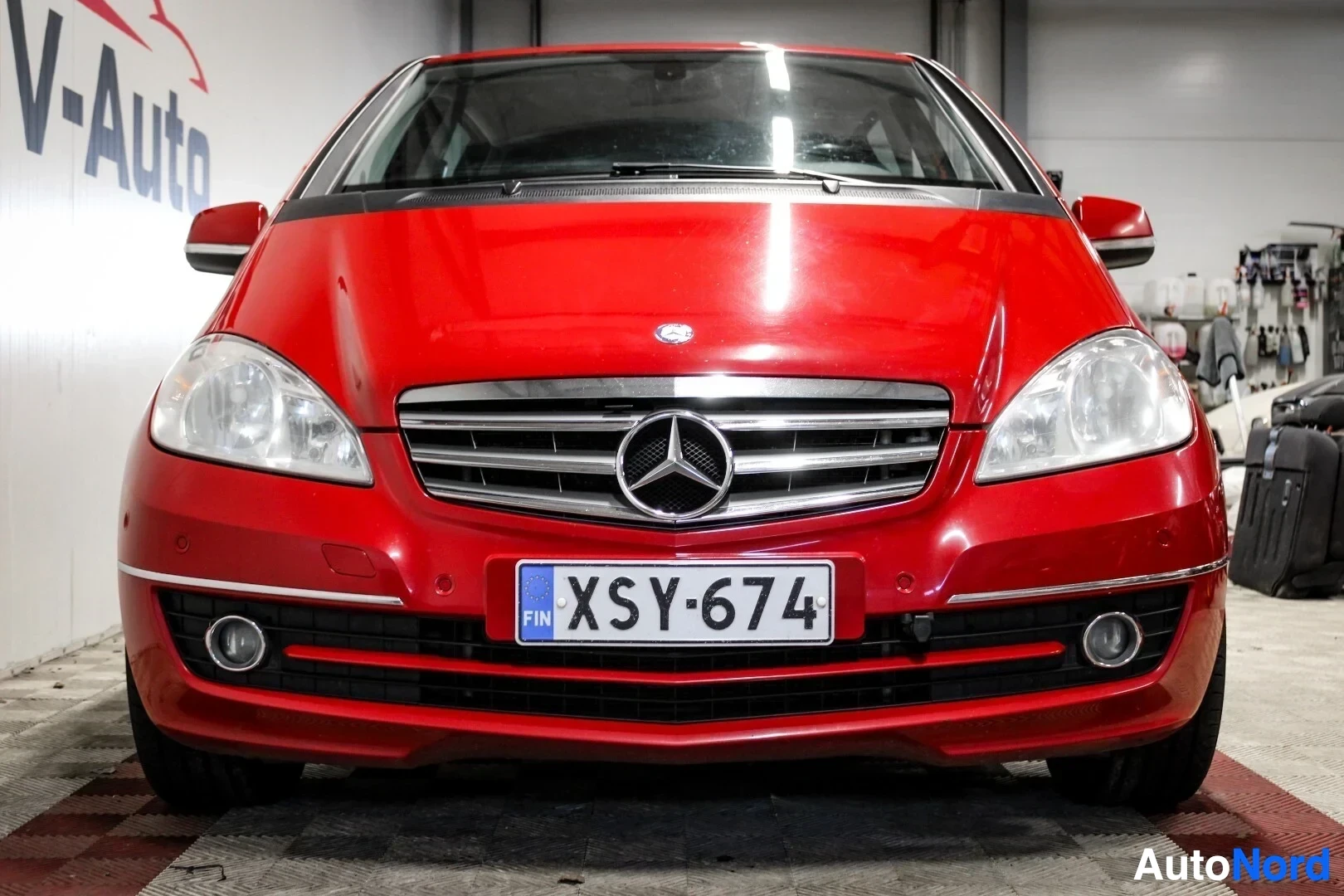 Edestä: Punaisen henkilöauton keula näkyy kuvassa. Autossa on kaksi pyöreää sumuvaloa etupuskurissa ja Mercedes-Benz-logo keskellä. Rekisterikilpi on ""