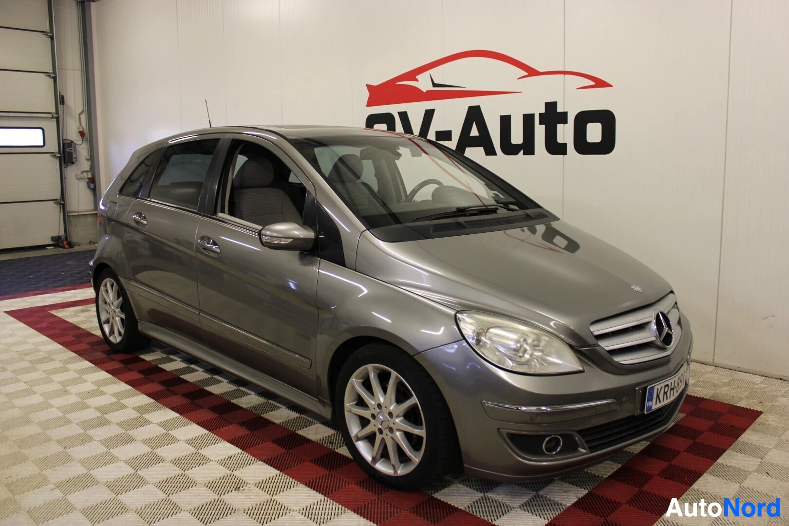 Etuviisto, nokka vasemmalle: Harmaa Mercedes-Benz B-sarja viistosti edestä päin kuvattuna autohallissa, OV-Auto-logo taustalla. Autossa on hopeat monipuolaiset kevytmetallivanteet