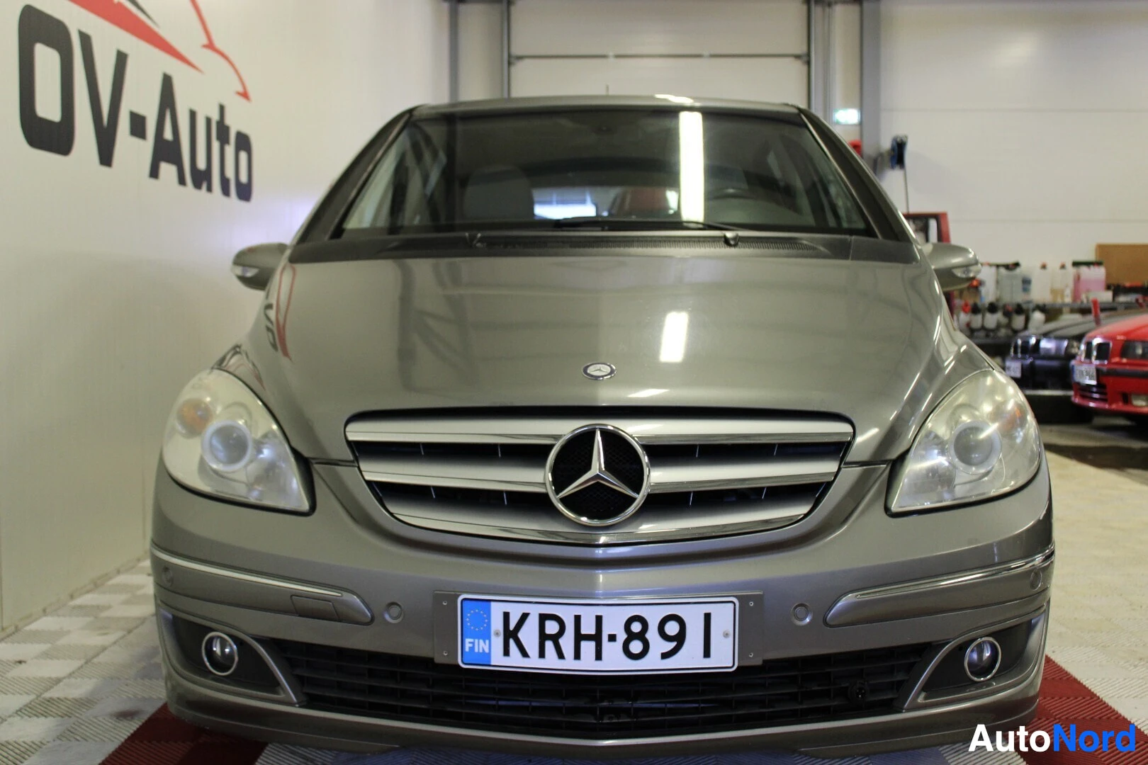 Edestä: Harmaa Mercedes-Benz B-sarja edestä päin kuvattuna autohallissa, OV-Auto-logo taustalla. Etuosassa on rekisterikilpi