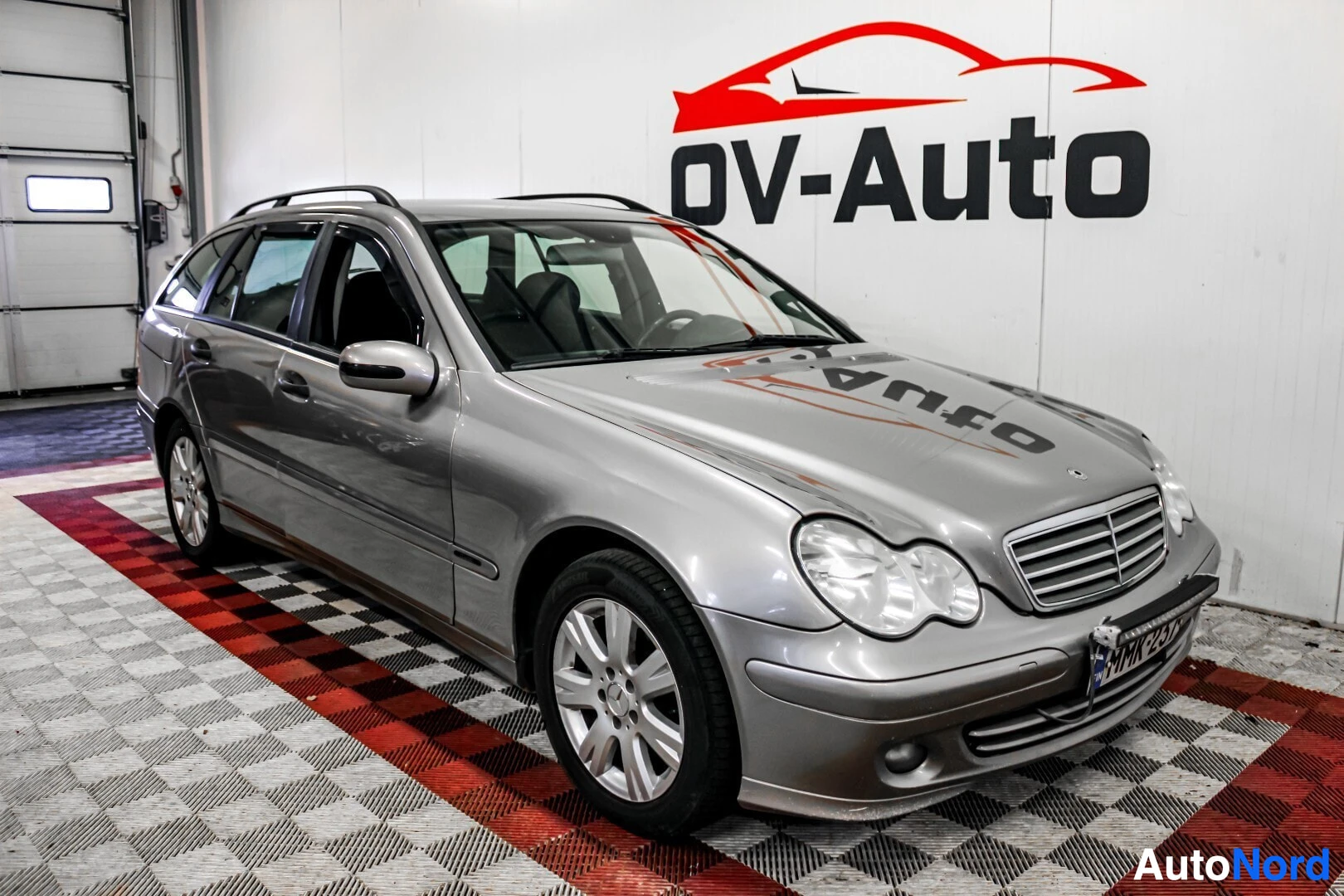 Etuviisto, nokka oikealle: Harmaa farmariauto edestä ja vasemmalta viistosti kuvattuna. Autossa on kattokaiteet ja alumiinivanteet. Taustalla näkyy OV-Auto-logo.