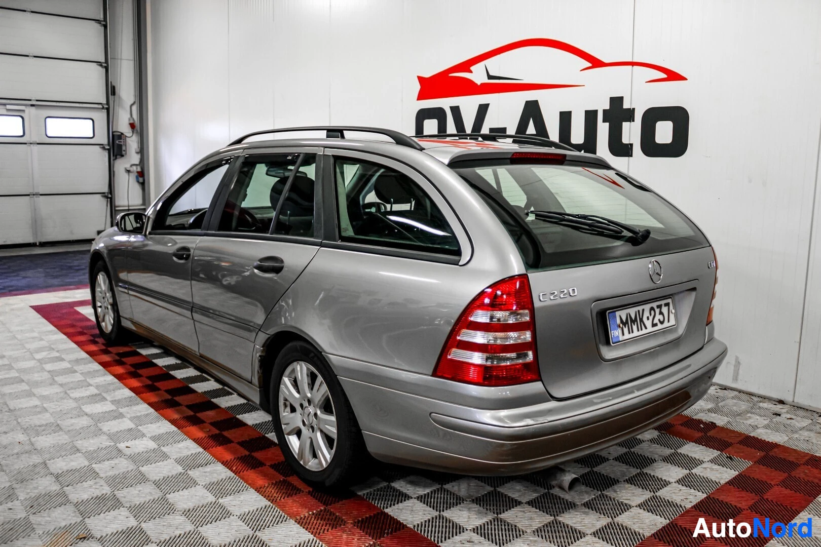 Takaviisto: Harmaa farmariauto takaa ja oikealta viistosti kuvattuna. Autossa on kattokaiteet ja takalasissa pyyhin. Takaluukussa näkyy mallimerkintä "C 220".