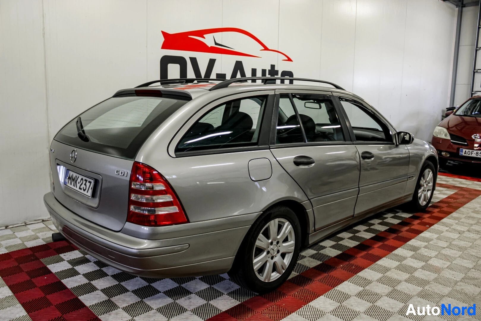 Takaviisto: Harmaa Mercedes-Benz C-sarjan farmariauto on kuvattu takaviistosta oikealta puolelta auton vasen kylki ja takaosa näkyvissä. Autossa on kattokiskot ja hopeiset