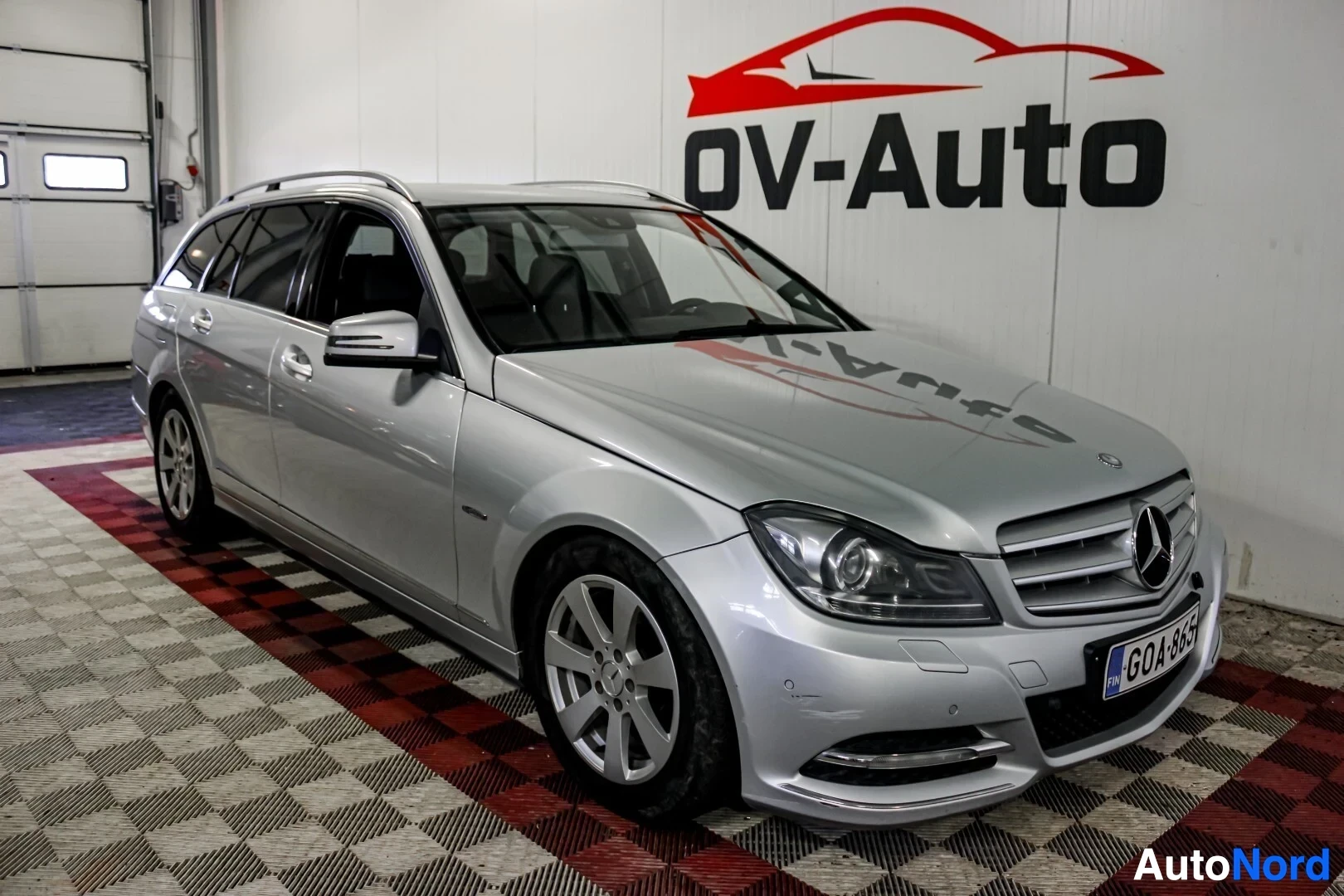 Etuviisto, nokka vasemmalle: Hopeanvärinen Mercedes-Benz-farmariauto kuvattuna etuviistosta oikealta, edessä kromattu maski ja suurikokoinen Mercedes-Benzin logo. Auton takaosassa