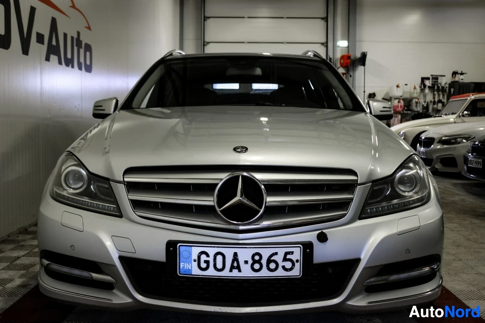 Edestä: Hopeanvärinen Mercedes-Benz-farmariauto kuvattuna edestä, edessä kromattu maski ja suurikokoinen Mercedes-Benzin logo. Ajovalot ovat päällä ja suunnattu eteenpä