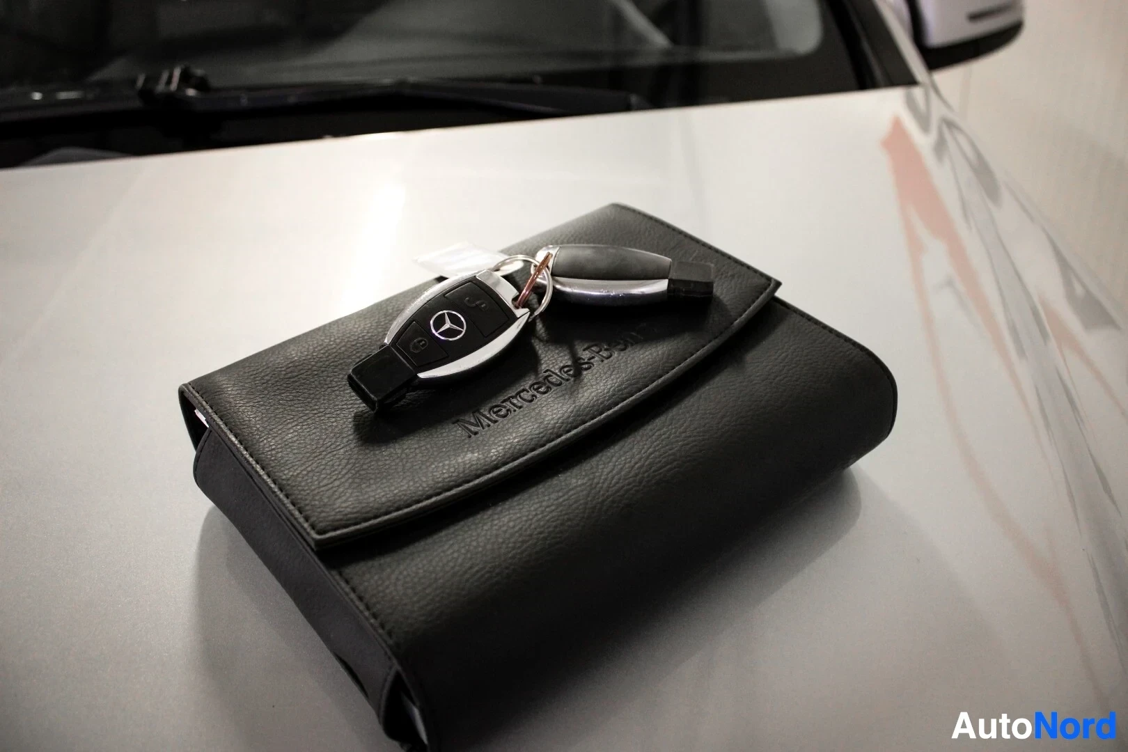 Yksityiskohta: Musta nahkainen auton käsikirjakotelo, jossa on "Mercedes-Benz" -logo, lepää ajoneuvon konepellillä. Kotelon päällä on kaksi mustaa auton avainta, joista toises