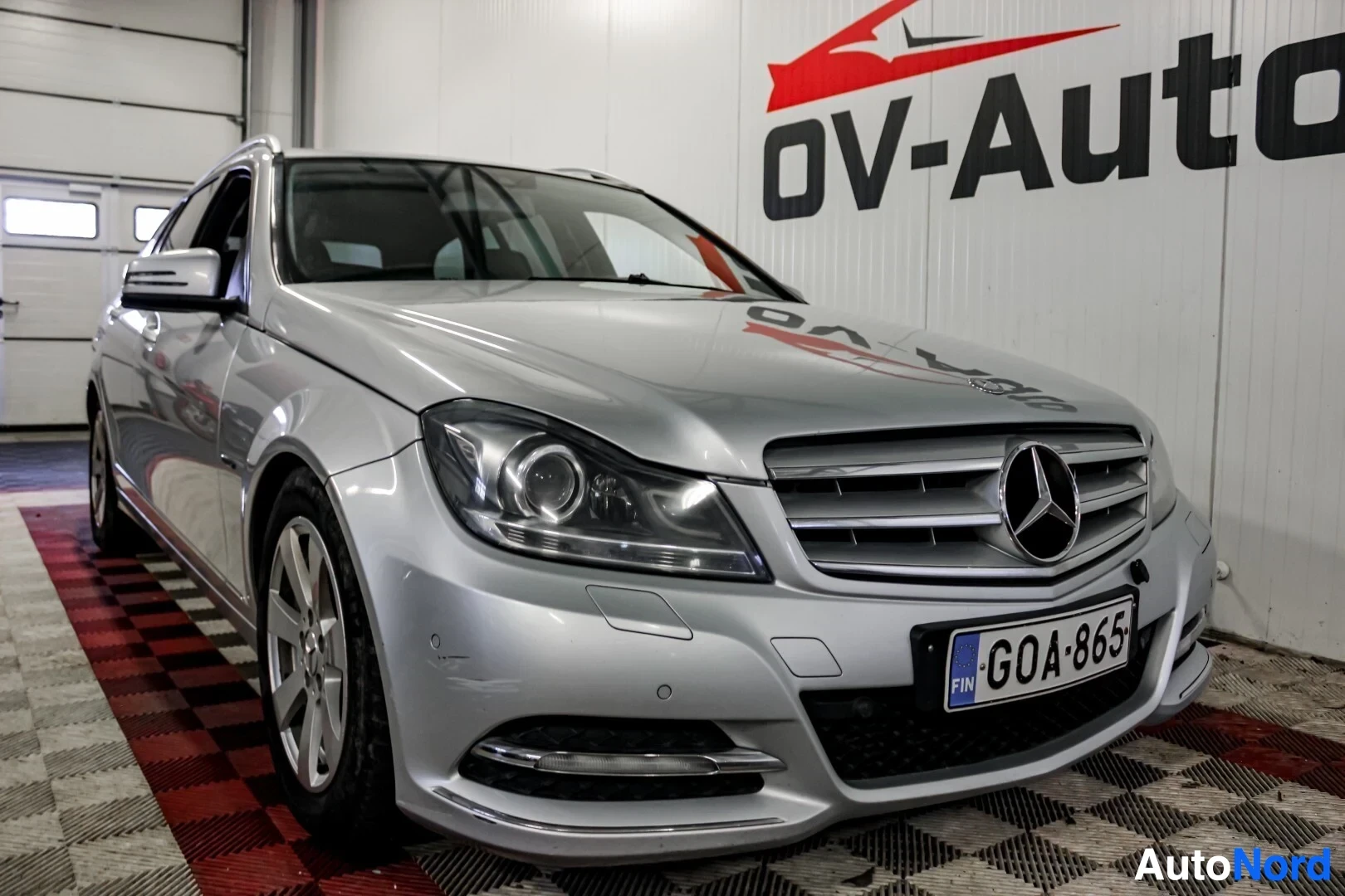 Etuviisto, nokka vasemmalle: Hopeanvärinen Mercedes-Benz-farmariauto kuvattuna etuviistosta vasemmalta, edessä kromattu maski ja suurikokoinen Mercedes-Benzin logo. Ajovalot on suu