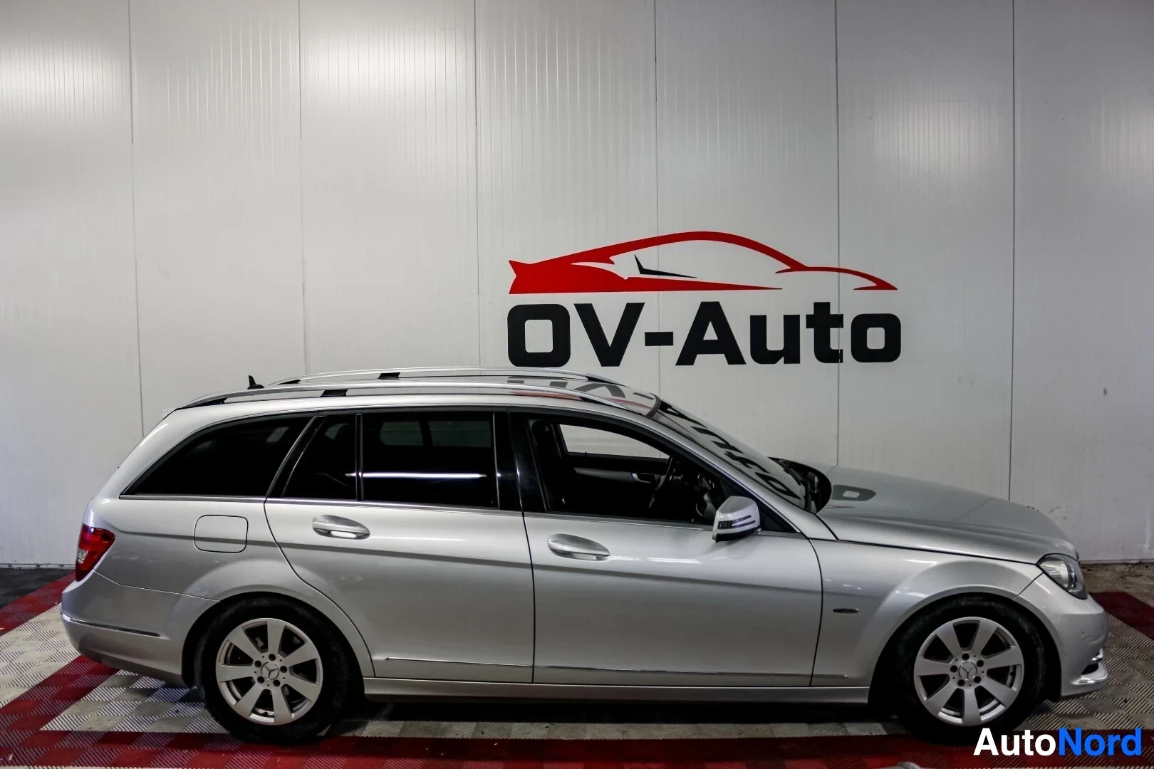 Sivulta: Hopeanvärinen Mercedes-Benz C-sarjan farmari, jossa on kattokaiteet ja viisispakkaiset kevytmetallivanteet. Auto on pysäköity ruudulliselle lattialle valkoisen