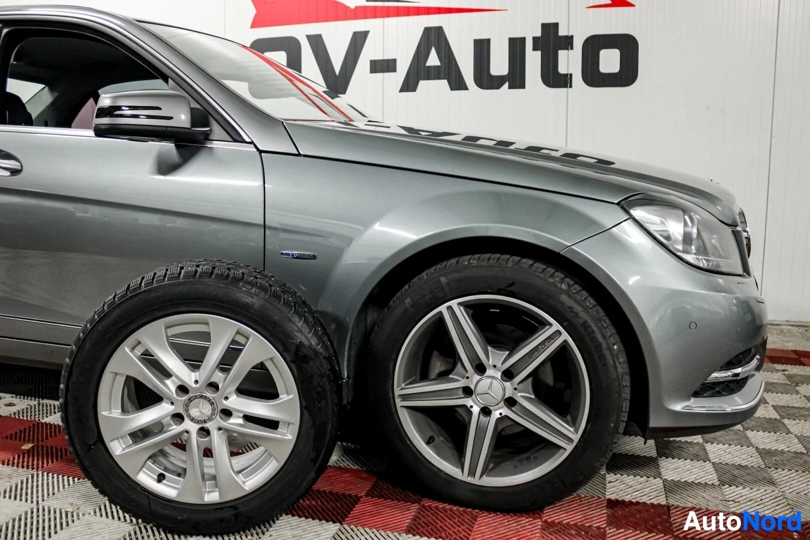 Etuviisto, nokka oikealle: Harmaa auto kuvattuna edestä oikealta puolelta, kaksi rengasta alumiinivanteineen on auton edessä maassa. Auton oikea eturengas on kiinni autossa ja sen
