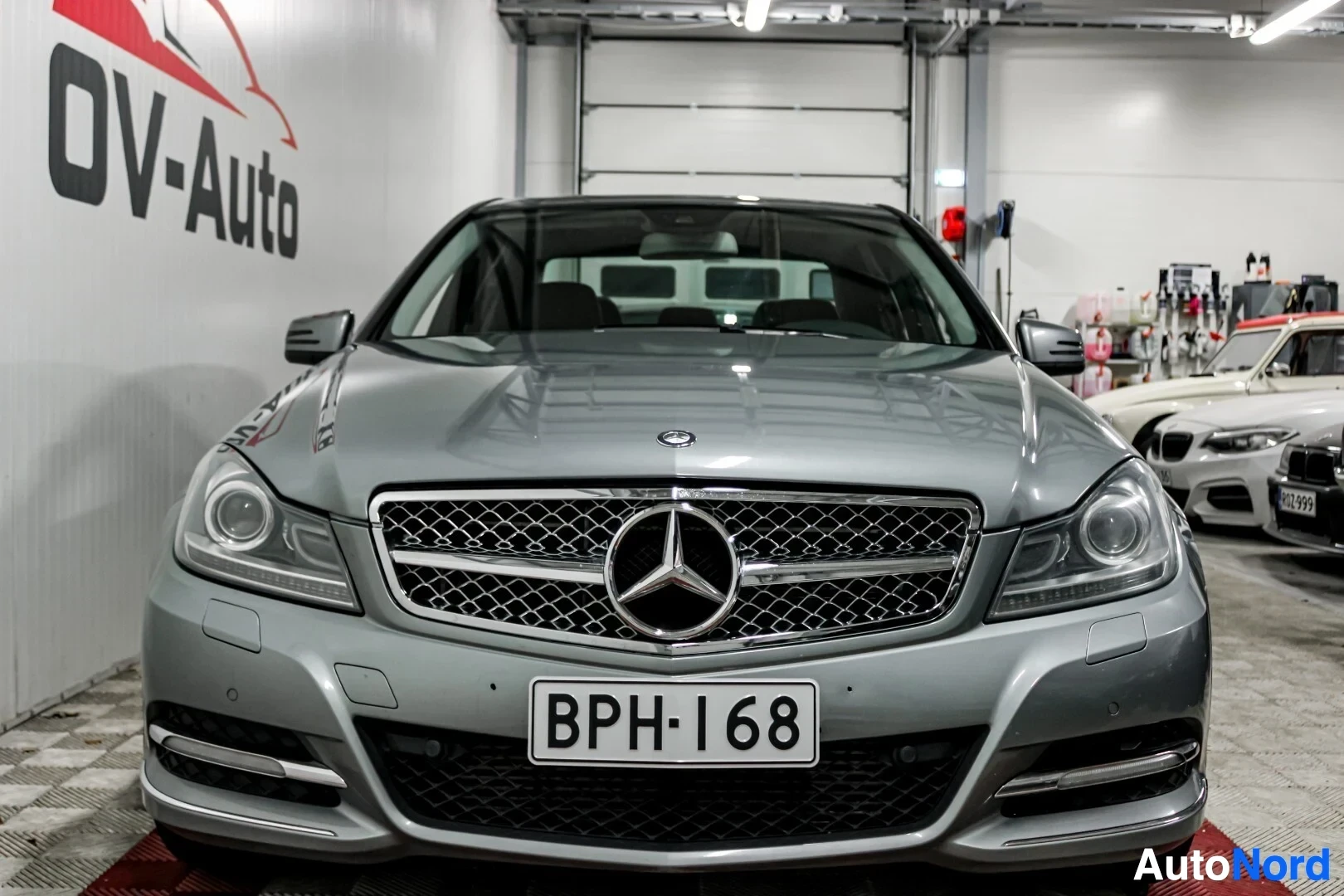 Edestä: Harmaan henkilöauton keula edestä päin. Rekisterikilvessä on teksti. Autossa on Mercedes-Benz-logo etusäleikössä. Valkoinen seinä, jossa on punamusta "O
