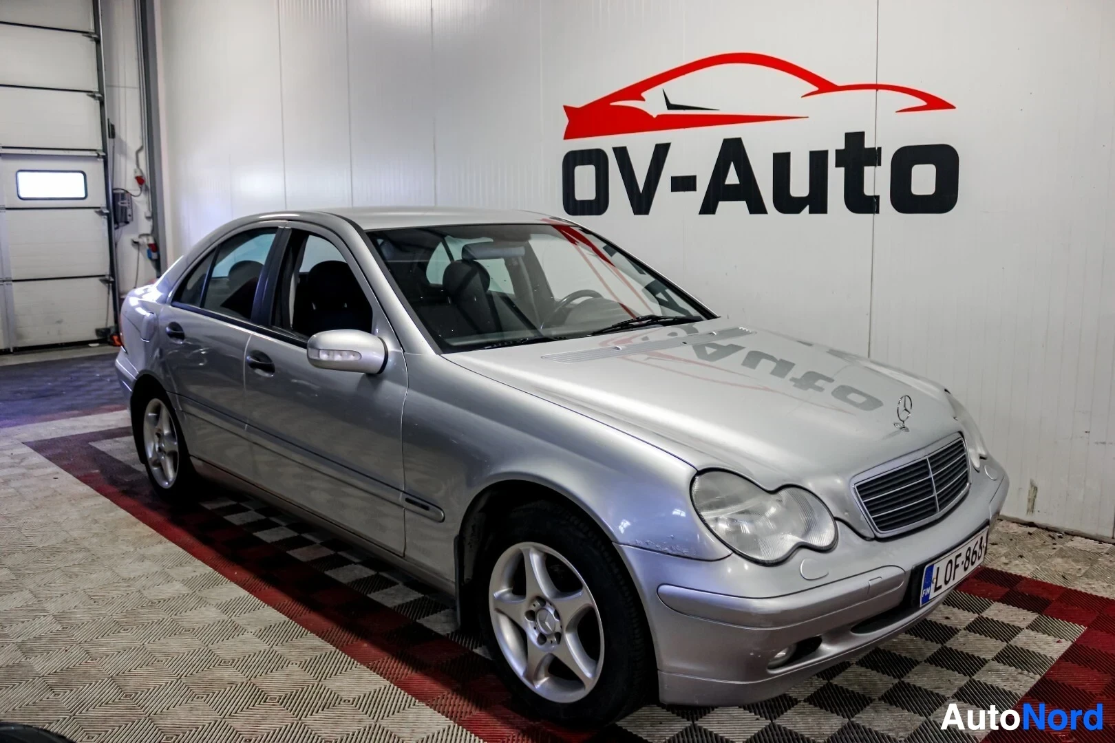 Etuviisto, nokka vasemmalle: Hopeanvärinen Mercedes-Benz C-sarja, rekisterinumero, kuvattuna edestä oikealta puolelta sisätiloissa. Auto on parkissa punaisen ja mustan ruut