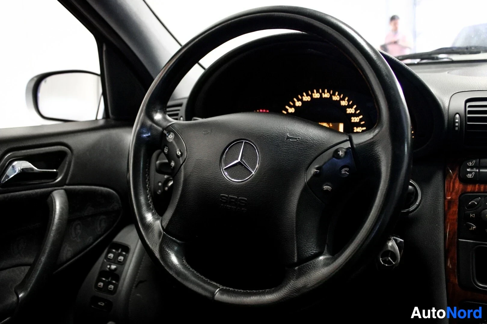 Yksityiskohta: Lähikuva ohjauspyörästä, jossa Mercedes-Benzin logo. Ohjauspyörässä on painikkeita ja mittaristossa näkyy oranssia taustavaloa.