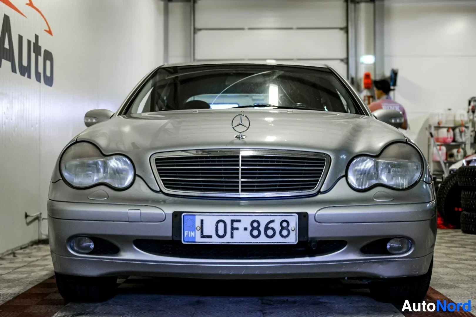 Edestä: Hopeanvärinen Mercedes-Benz C-sarja, rekisterinumero, kuvattuna suoraan edestä sisätiloissa. Auto on parkissa valkoisen seinän edessä ja taustalla näkyy