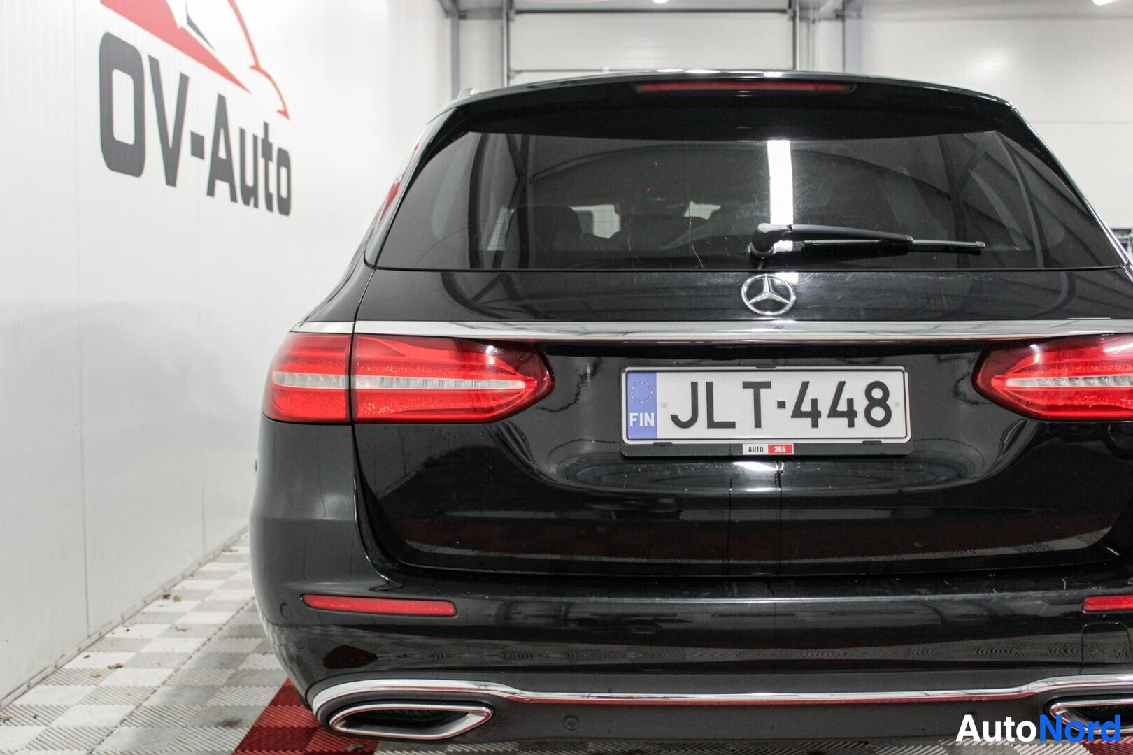 Takaa: Auton takaosa, jossa näkyy rekisterikilpi "" ja Mercedes-Benz-logo. Auto on pysäköity ruudulliselle lattialle sisätiloissa