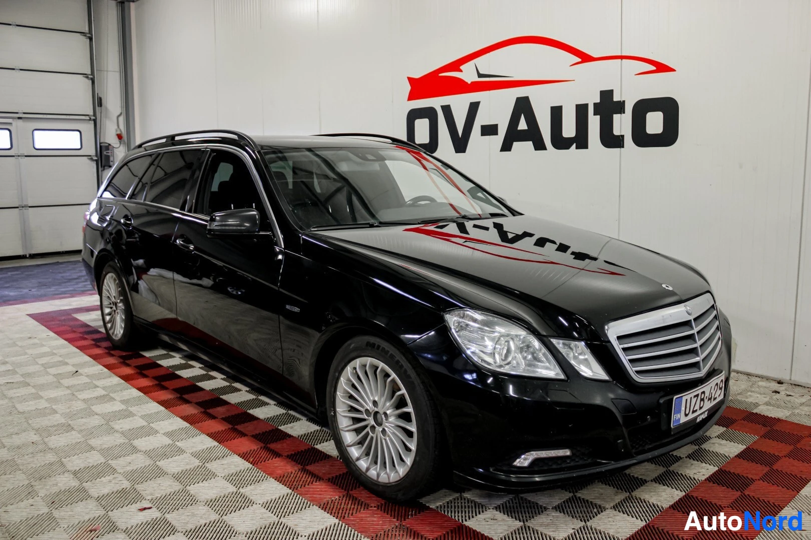 Etuviisto, nokka oikealle: Musta Mercedes-Benz E-sarjan farmariauto on pysäköity sisätiloihin, edestä ja vasemmalta sivulta kuvattuna. Autossa on hopeanväriset monipuolaiset kevytm
