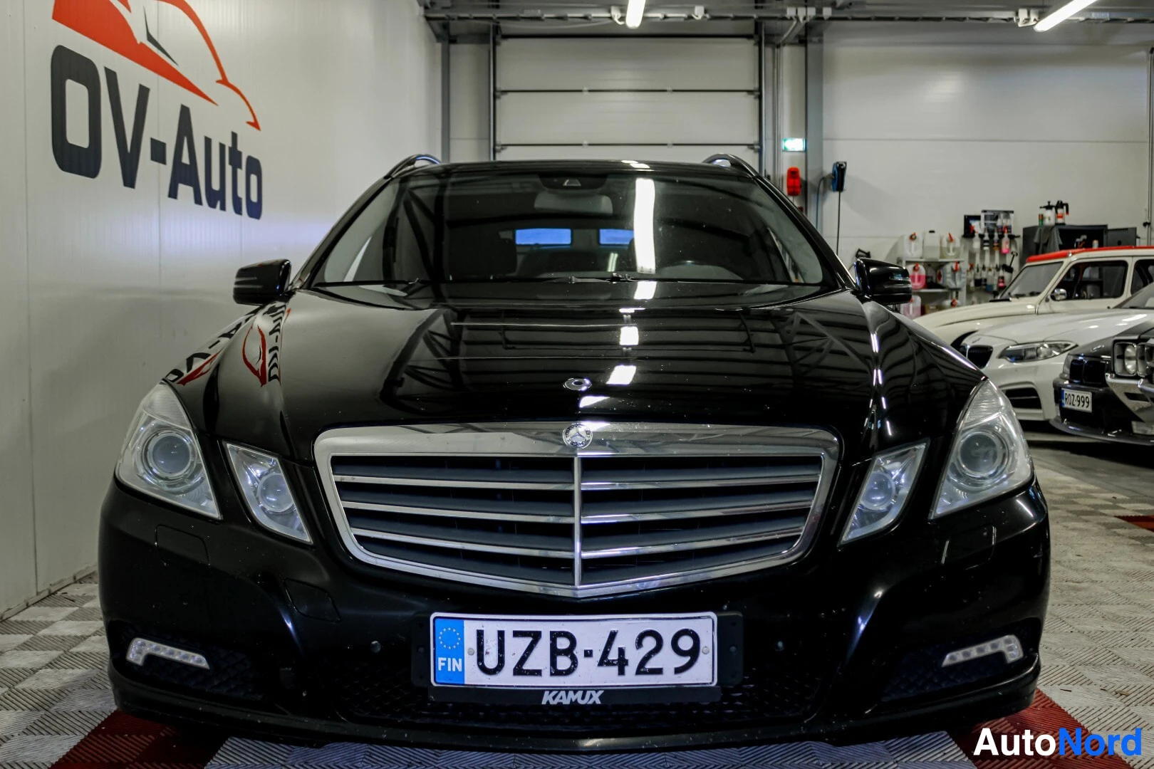 Edestä: Musta Mercedes-Benz E-sarjan farmariauto edestä päin kuvattuna sisätiloissa. Autossa on kromattu etusäleikkö ja etupuskurissa päiväajovalot. Rekisterikilpi UZB-
