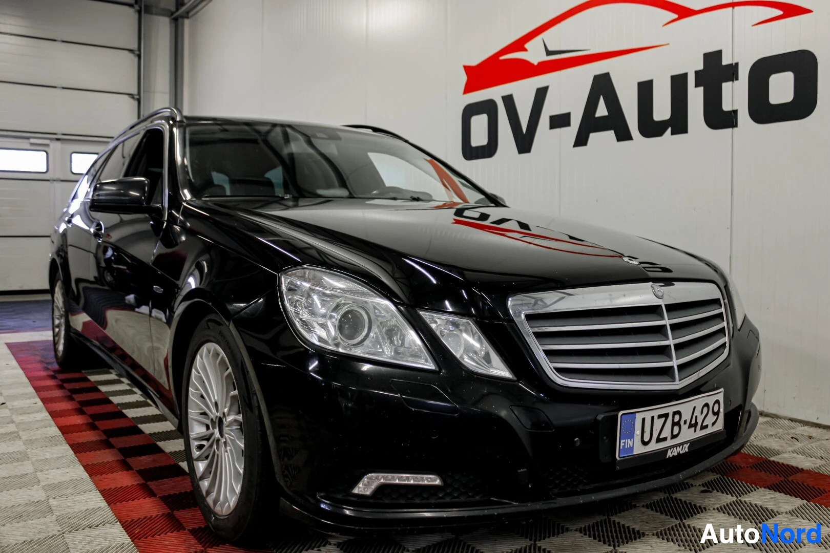 Etuviisto, nokka oikealle: Musta Mercedes-Benz E-sarjan farmariauto on pysäköity sisätiloihin, edestä ja vasemmalta sivulta kuvattuna. Autossa on hopeanväriset monipuolaiset kevytm