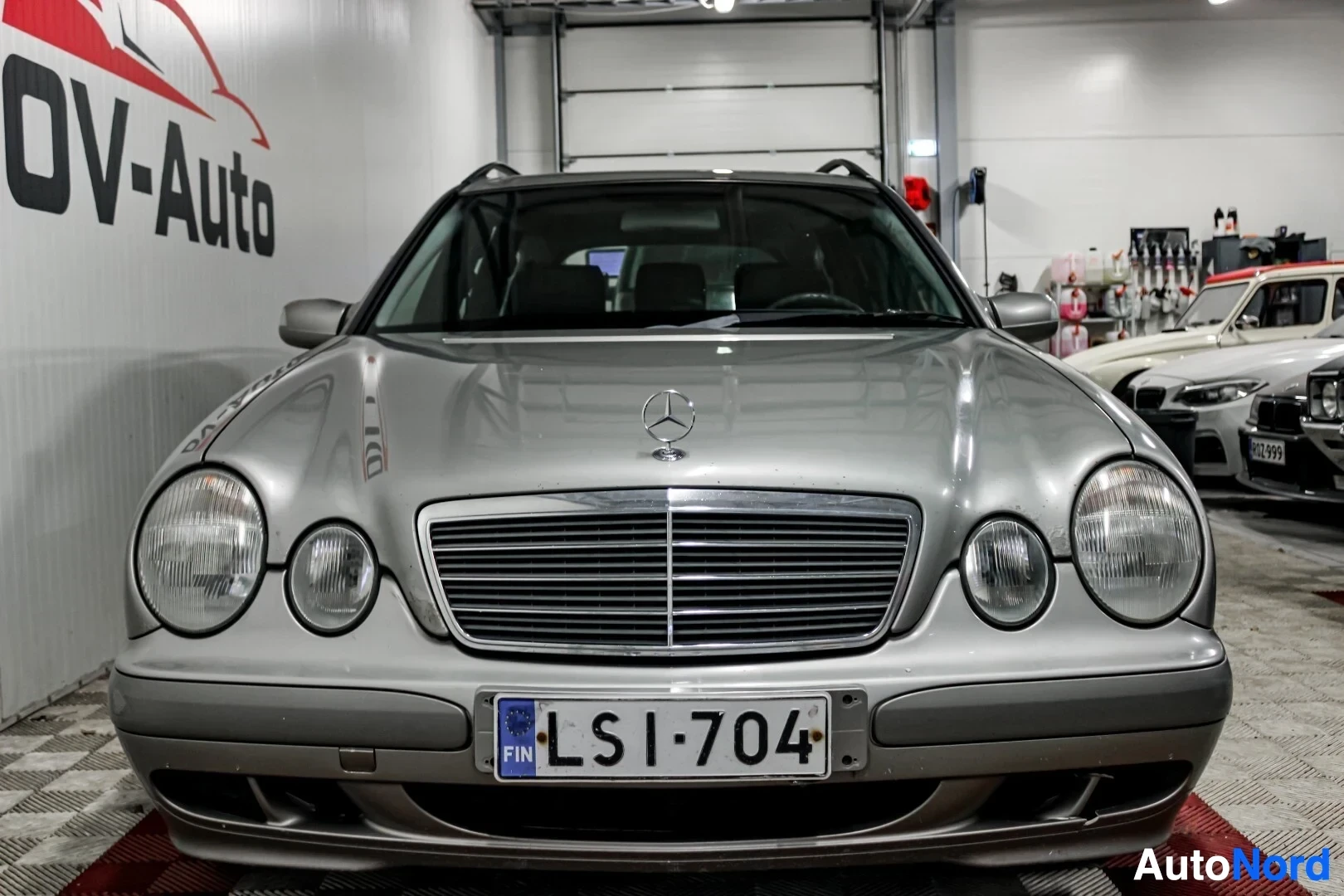 Edestä: Harmaan farmariauton etuosa, jossa on Mercedes-Benz-logo ja avoin säleikkö. Eturekisterikilpi "" on näkyvissä