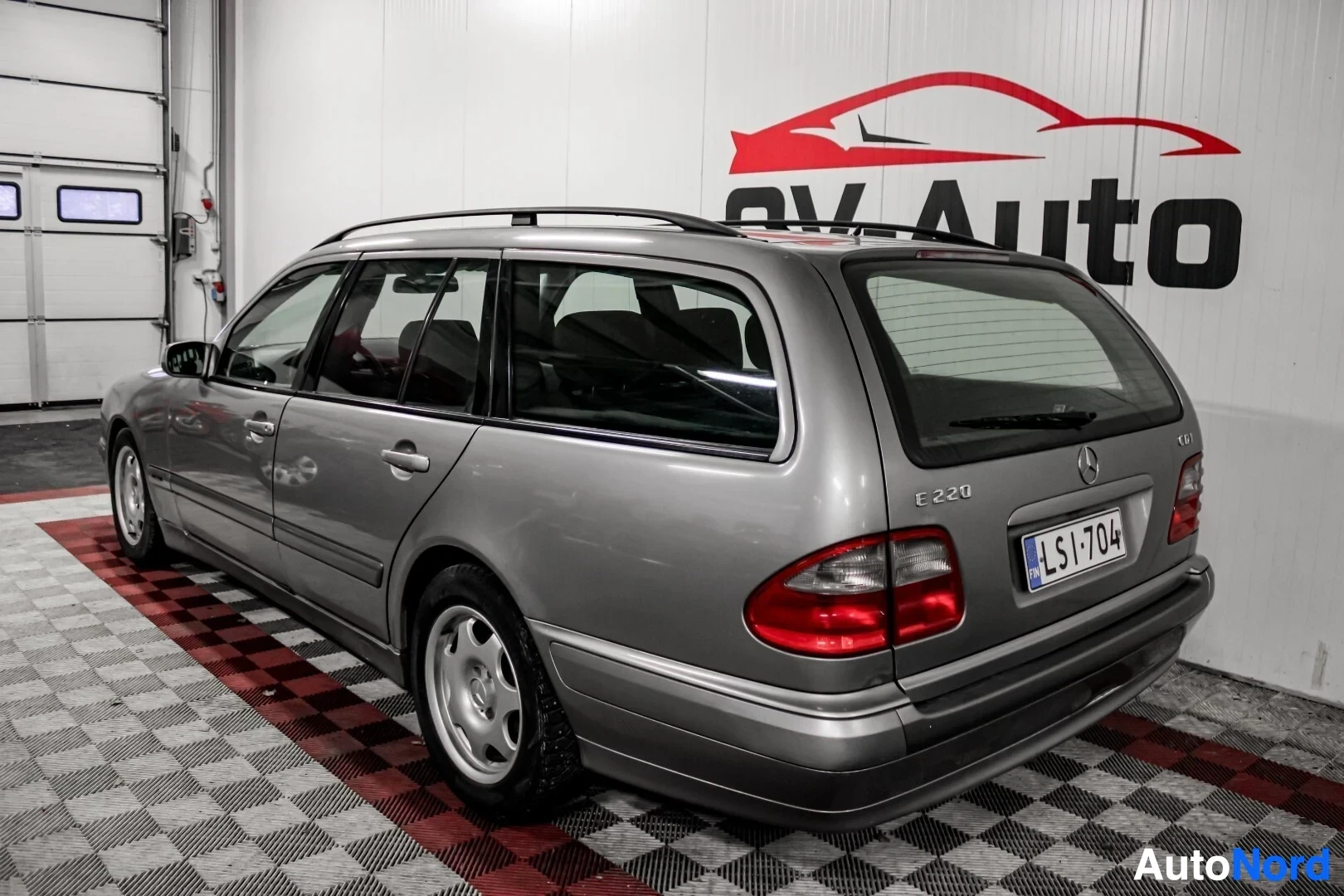 Takaviisto: Harmaa farmariauto, jossa on kattokaiteet ja kevytmetallivanteet, kuvattuna takaa oikealta viistosta sisätilassa. Takarekisterikilpi "" ja "E 220" -merki