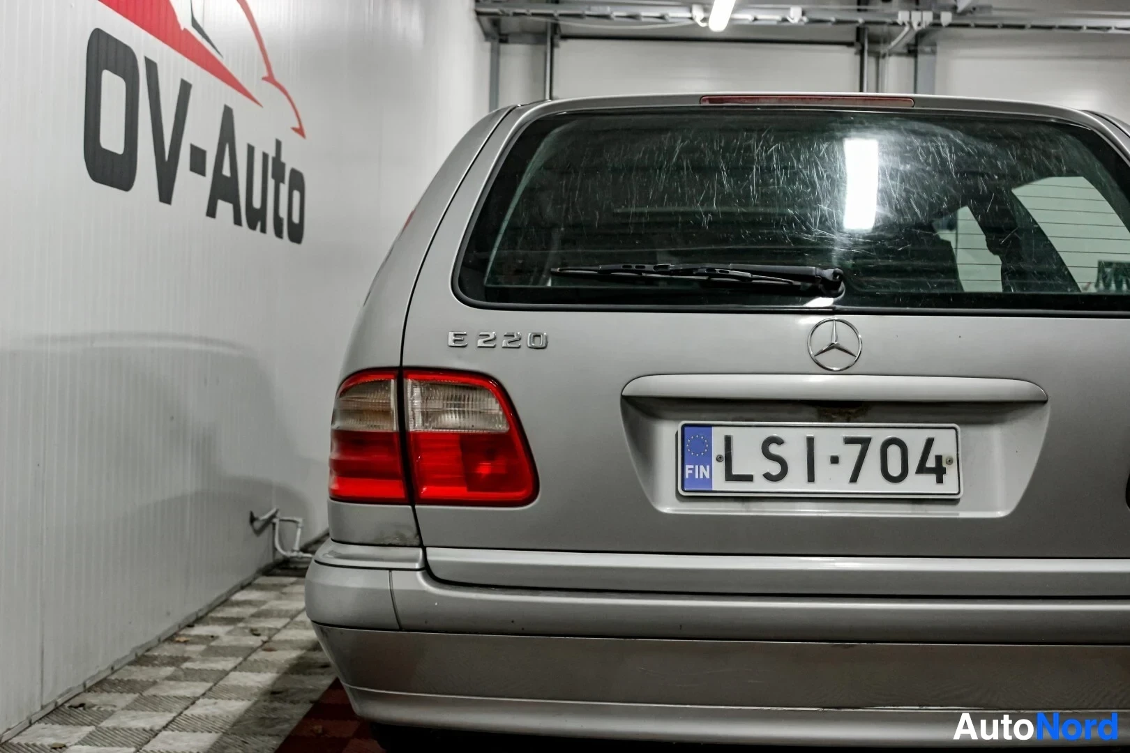Takaa: Hopeanvärinen farmariauto takaapäin kuvattuna. Takaosassa näkyy Mercedes-Benz-logo, E 220 -merkintä ja rekisterikilpi