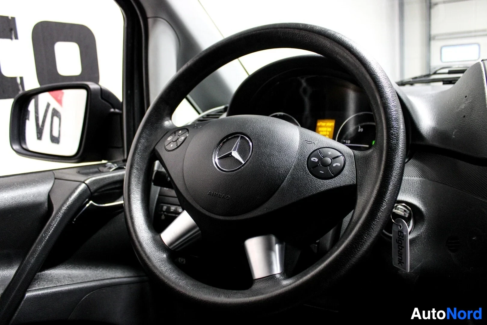 Ohjaamo: Lähikuva ohjauspyörästä, jossa on Mercedes-Benzin logo ja monitoiminäppäimet. Virtalukossa roikkuu avaimet. Vasemmalla näkyy auton ulkopeili.