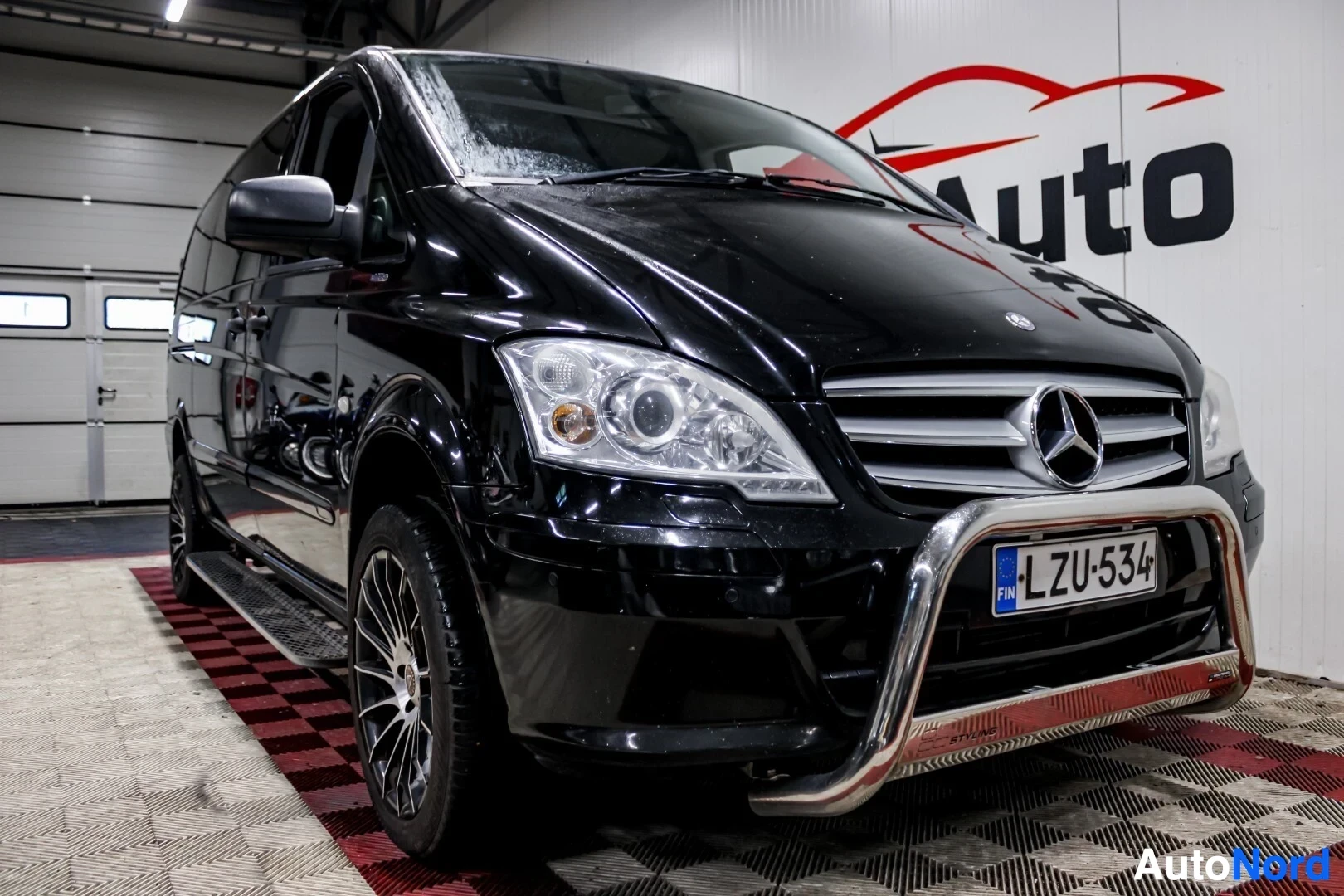Etuviisto, nokka oikealle: Musta Mercedes-Benz Vito -pakettiauto kuvattuna edestä ja oikealta edestä, tarkempi kuva. Ajoneuvossa on krominen puskurinsuojus ja monipuolaiset kevytme