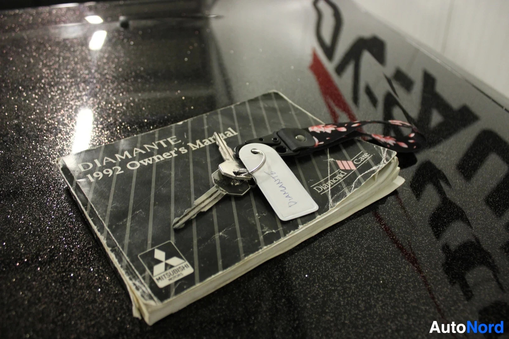 Yksityiskohta: Lähikuva auton avaimista, jotka makaavat "DIAMANTE 1992 Owner's Manual" -nimisen käyttöoppaan päällä. Kansi on musta ja siinä on valkoinen Mitsubishi-logo. Avai