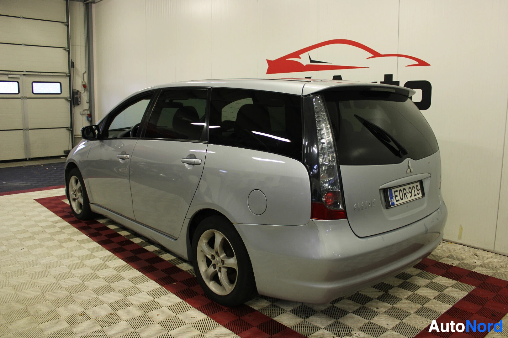 Takaviisto: Hopeanvärinen tila-auto kuvattuna takaviistosta oikealta puolelta sisätiloissa. Auton takana ja oikealla puolella näkyy ruudullinen lattia. Auton rekisterikilve