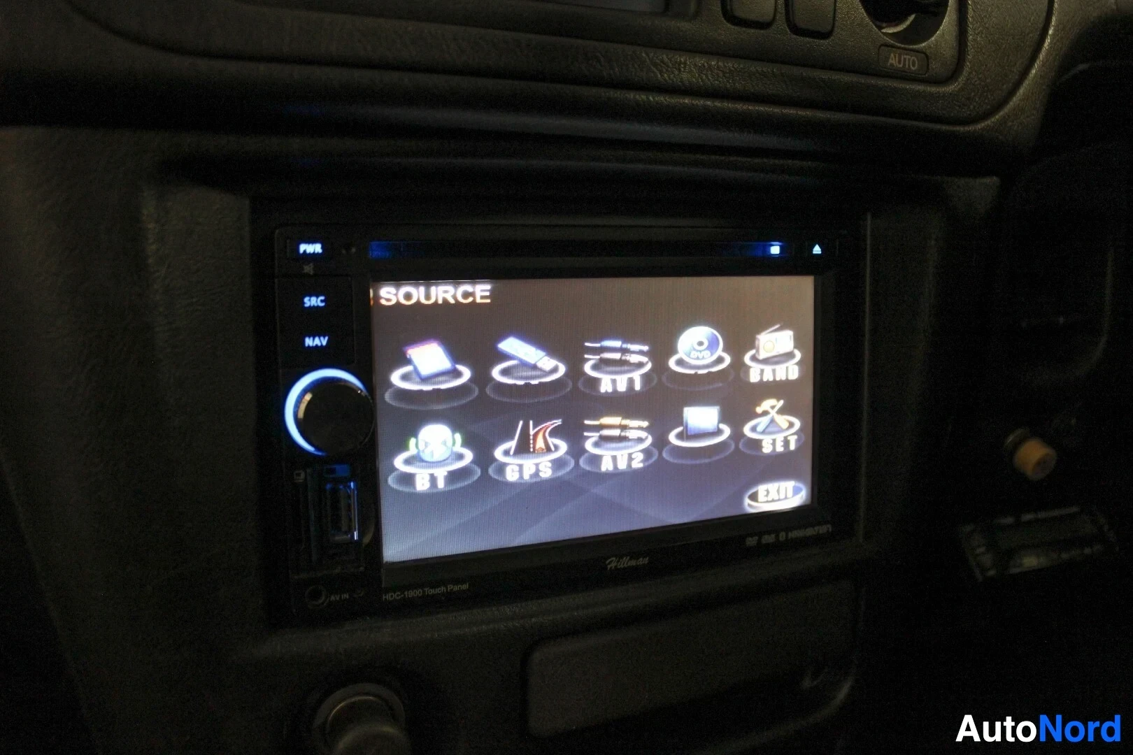 Ohjaamo: Lähikuva auton sisätilasta, jossa näkyy auton infotainment-järjestelmän näyttö. Näytöllä on useita kuvakkeita, kuten "SOURCE", "BT", "GPS", "AV1", "AV2" ja "EXI