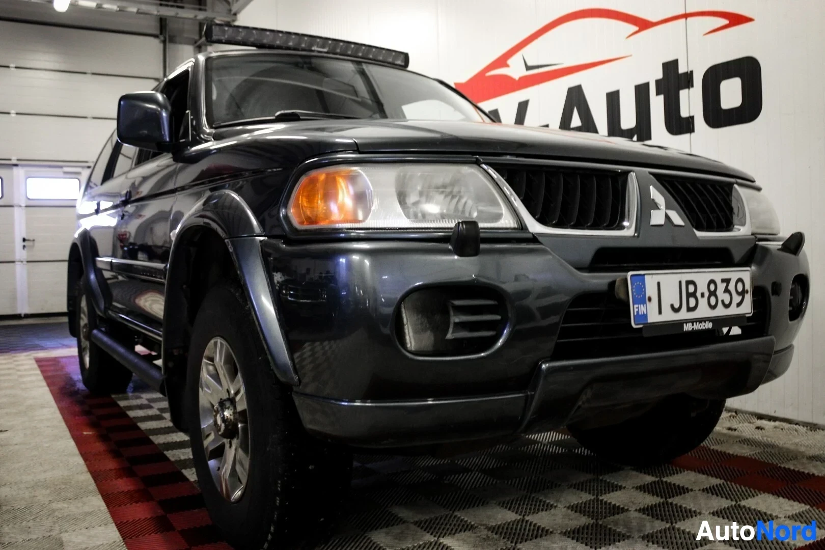 Etuviisto, nokka vasemmalle: Tummanharmaa Mitsubishi Pajero Sport -maastoauto kuvattuna matalasta kulmasta edestä, vasemmalta viistosta. Autossa on valopalkki katolla, astinlaudat