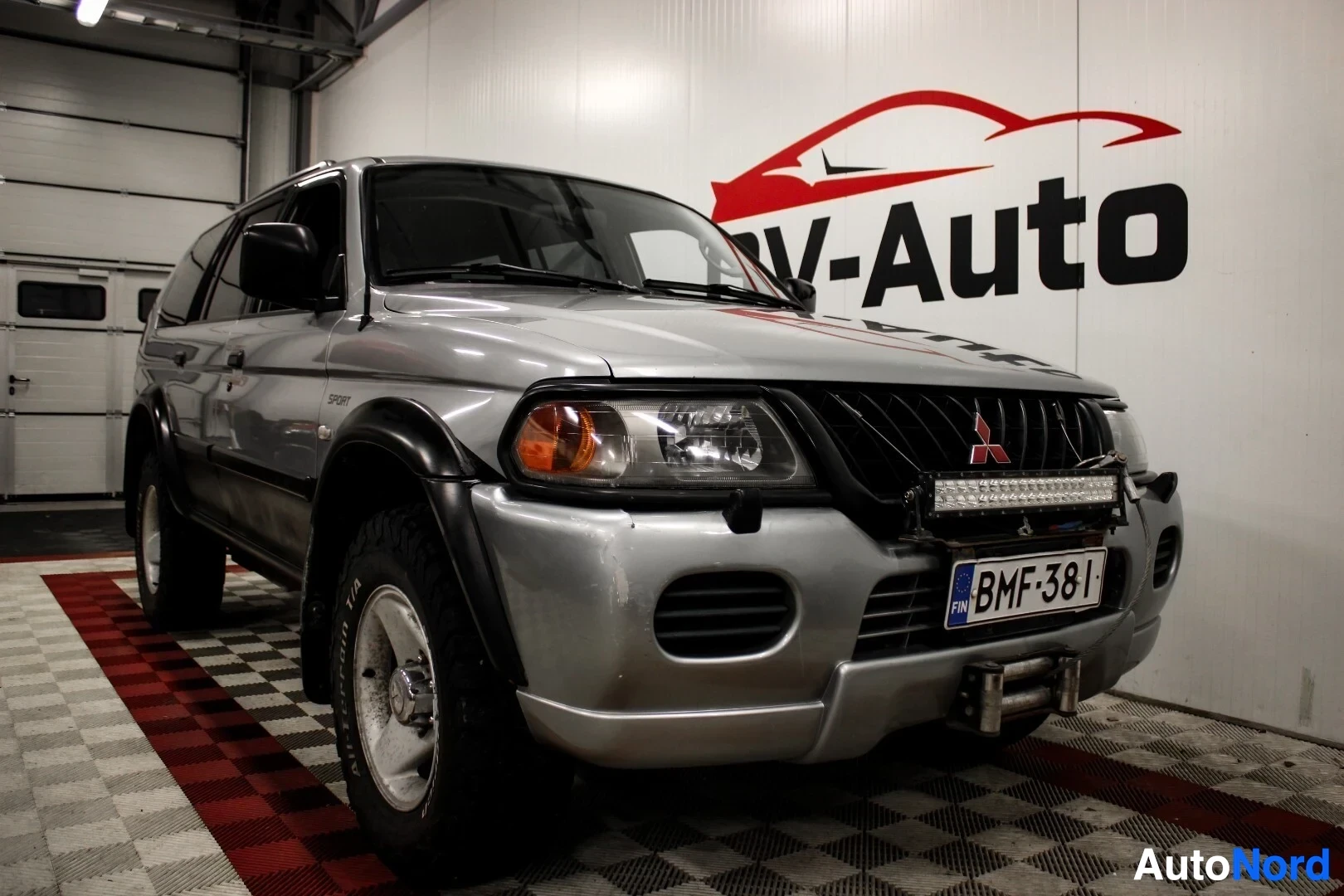 Etuviisto, nokka vasemmalle: Hopeanvärinen Mitsubishi Pajero Sport edestä ja oikealta sivulta kuvattuna sisätiloissa. Autossa on kattokaiteet, mustat pyöränkaaret ja keulassa lisäv