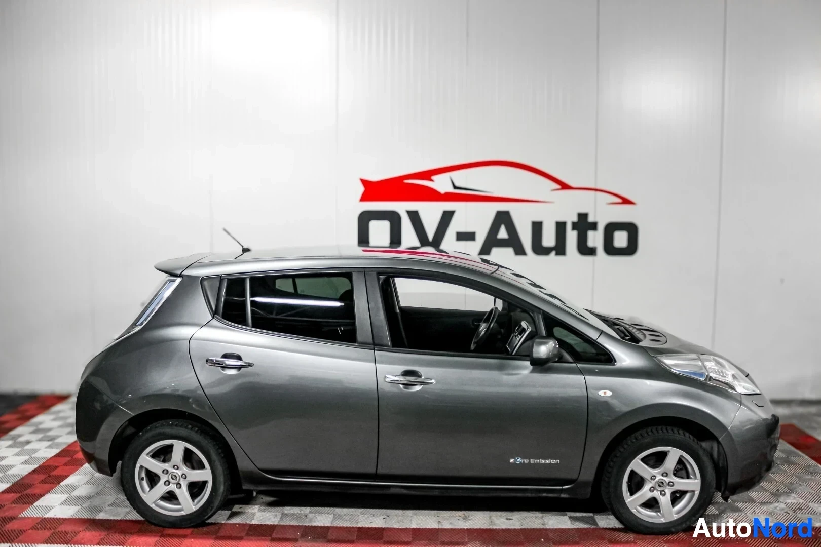 Sivulta: Hopeanharmaa Nissan Leaf autonäyttelytilassa, kuvattuna suoraan vasemmalta kyljeltä. Ajoneuvon vasemmassa kyljessä on 'e-power' tai vastaava merkintä etuoven ta