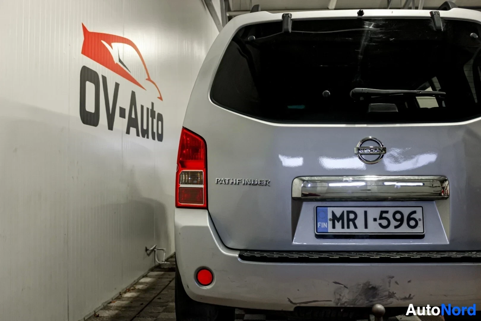 Takaa: Nissan Pathfinderin peräosan lähikuva, jossa näkyy vasen takavalo, Nissan-logo, Pathfinder-merkintä ja rekisterikilpi MR I-596. Seinällä on OV-Auto-logo.