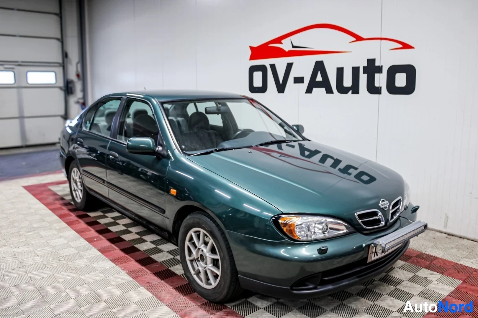 Etuviisto, nokka oikealle: Tummanvihreä henkilöauto pysäköitynä valkoisen seinän eteen, jossa on punamusta "OV-Auto"-logo. Auton edessä ja kyljessä on valkoisia ja punaisia ruudull