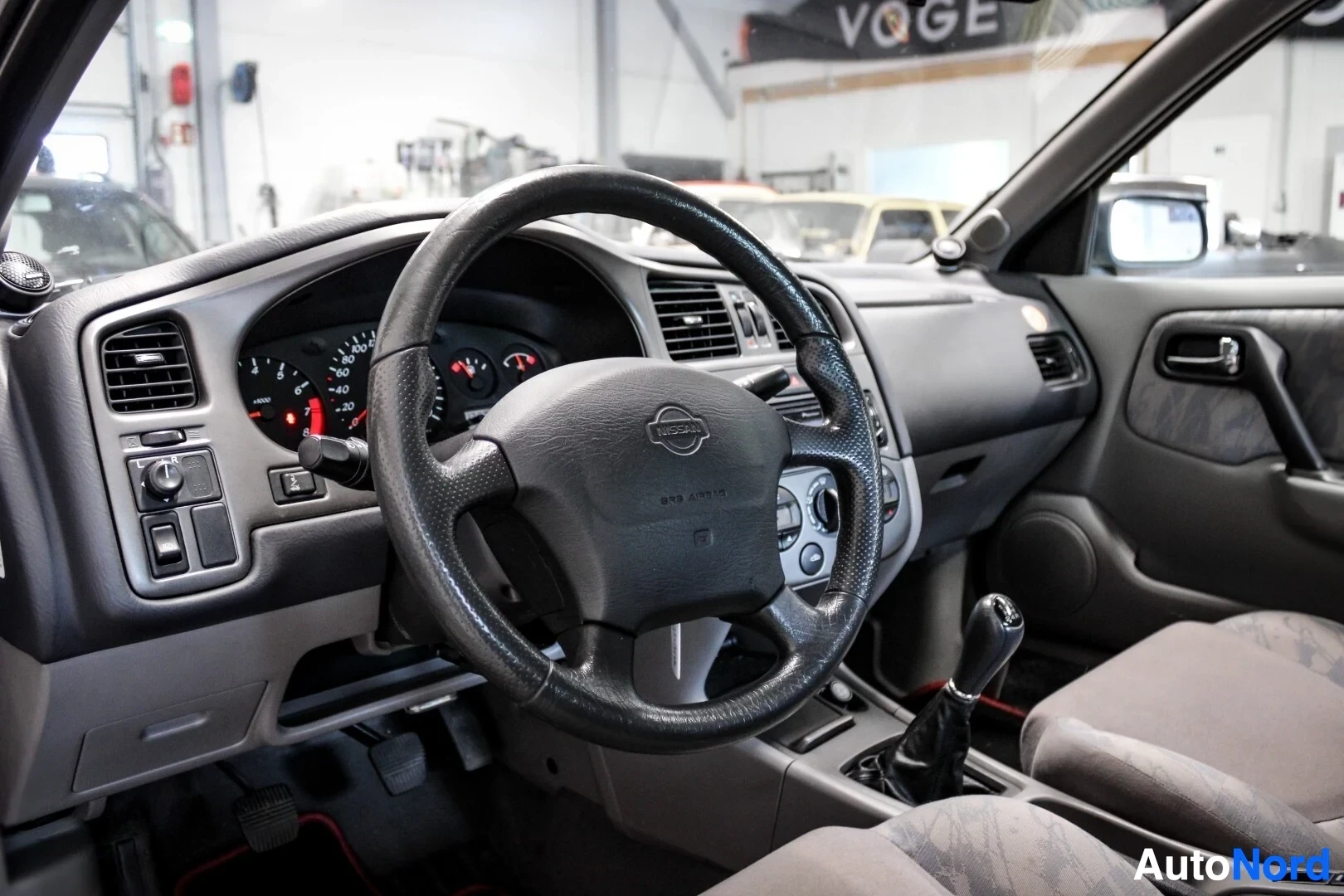Yksityiskohta: Lähikuva auton ohjaamosta kuljettajan puolelta, näyttäen ohjauspyörän, kojelaudan ja vaihdekepin. Ohjauspyörässä on Nissan-logo. Etuistuin näkyy oikeassa alakul