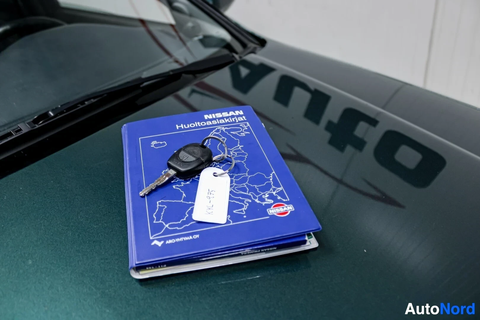 Yksityiskohta: Avaimet ja sininen huoltokirja kannen tekstillä "NISSAN Huoltoasiakirjat" auton konepellillä. Avaimissa valkoinen avaimenperä, jossa ""