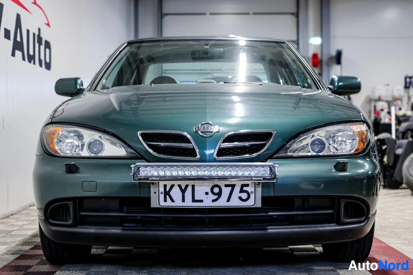 Edestä: Tummanvihreän henkilöauton etuosa suoraan edestäpäin. Etusäleikössä on Nissan-logo. Etupuskurissa on rekisterikilpi "" ja sen yläpuolella pitkä LED-valop