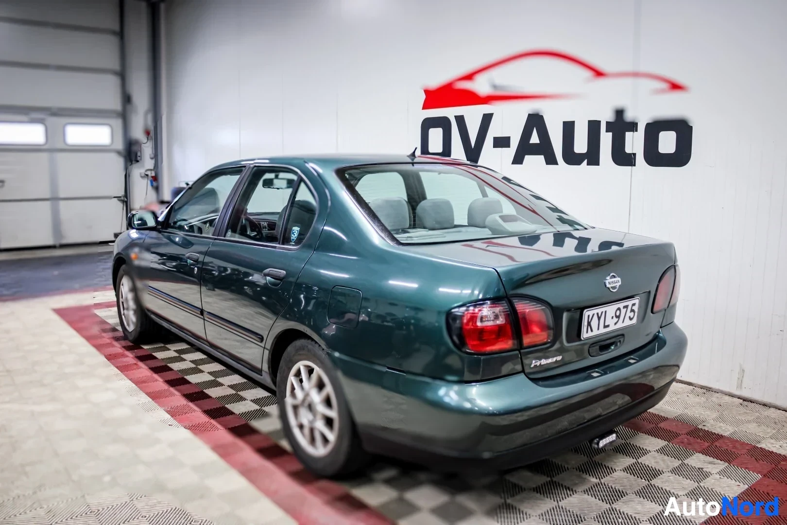 Takaviisto: Tummanvihreä henkilöauto kuvattuna takavasemmalta. Auton perässä on vetokoukku ja rekisterikilpi. Valkoisella seinällä takana on "OV-Auto"-logo.