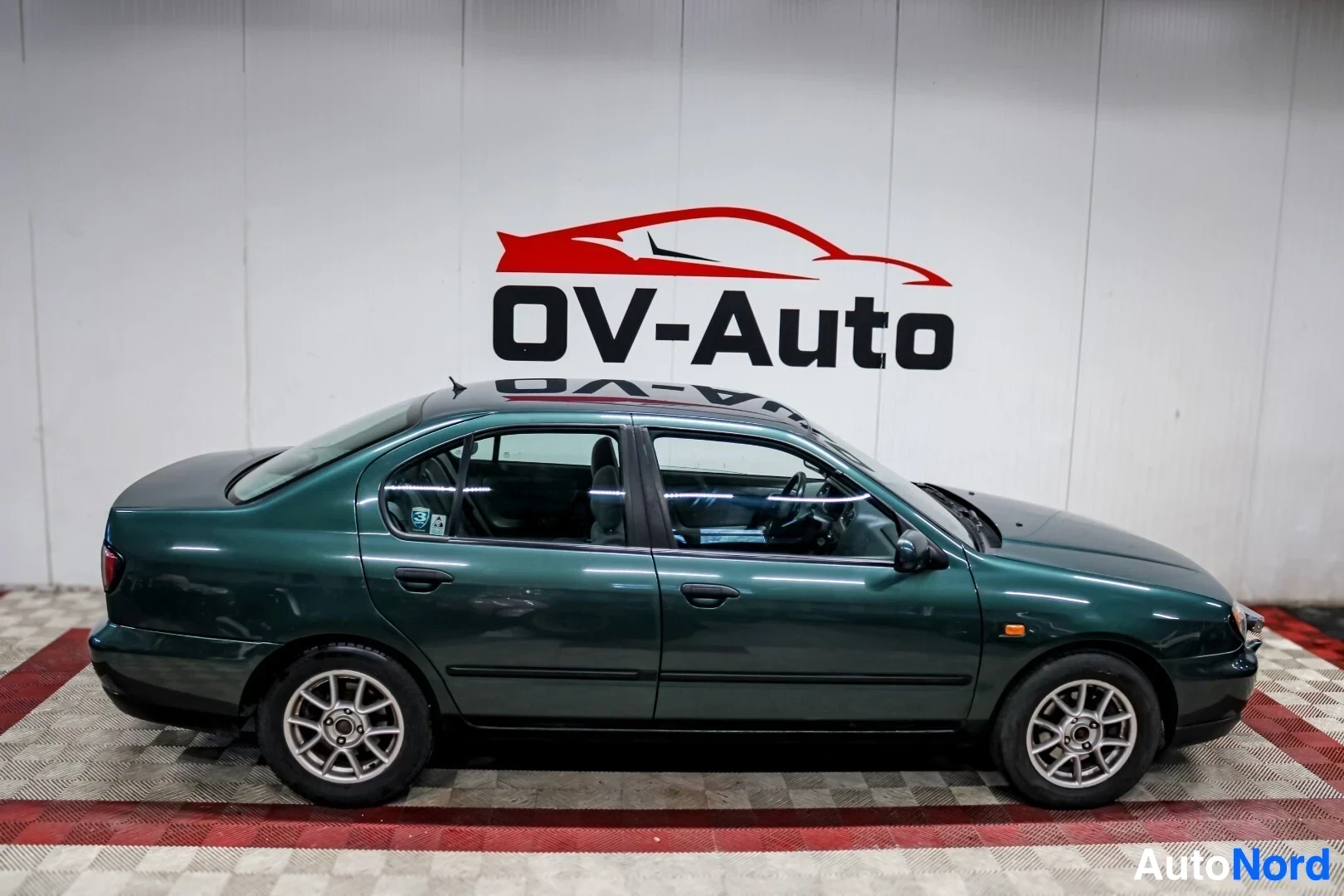 Sivulta: Tummanvihreä henkilöauto kuvattuna sivusta. Auto on parkissa valkoisessa hallissa, jonka seinällä on punainen ja musta OV-Auto-logo. Autossa on viisipuolaiset k