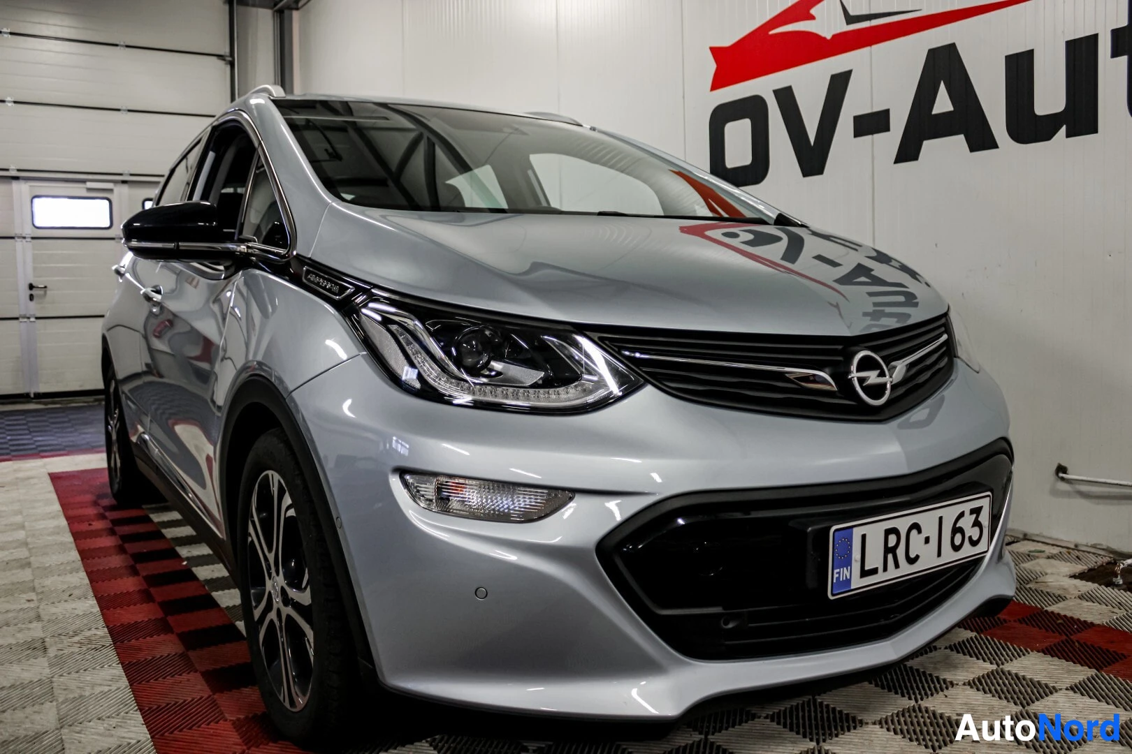 Etuviisto, nokka oikealle: Hopeanvärinen Opel Ampera-e etuviistosta edessä ja oikealla, ajovalot ja etupuskuri näkyvissä. Eturekisterikilpi on näkyvillä